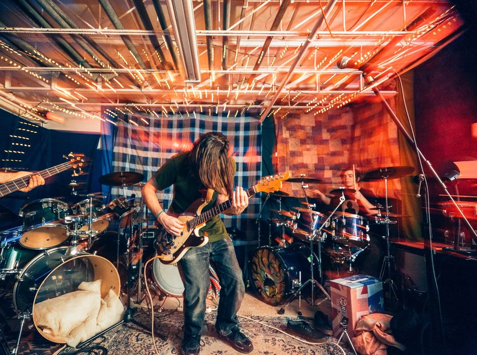 A man is playing a guitar in front of a drum set in a dark room.