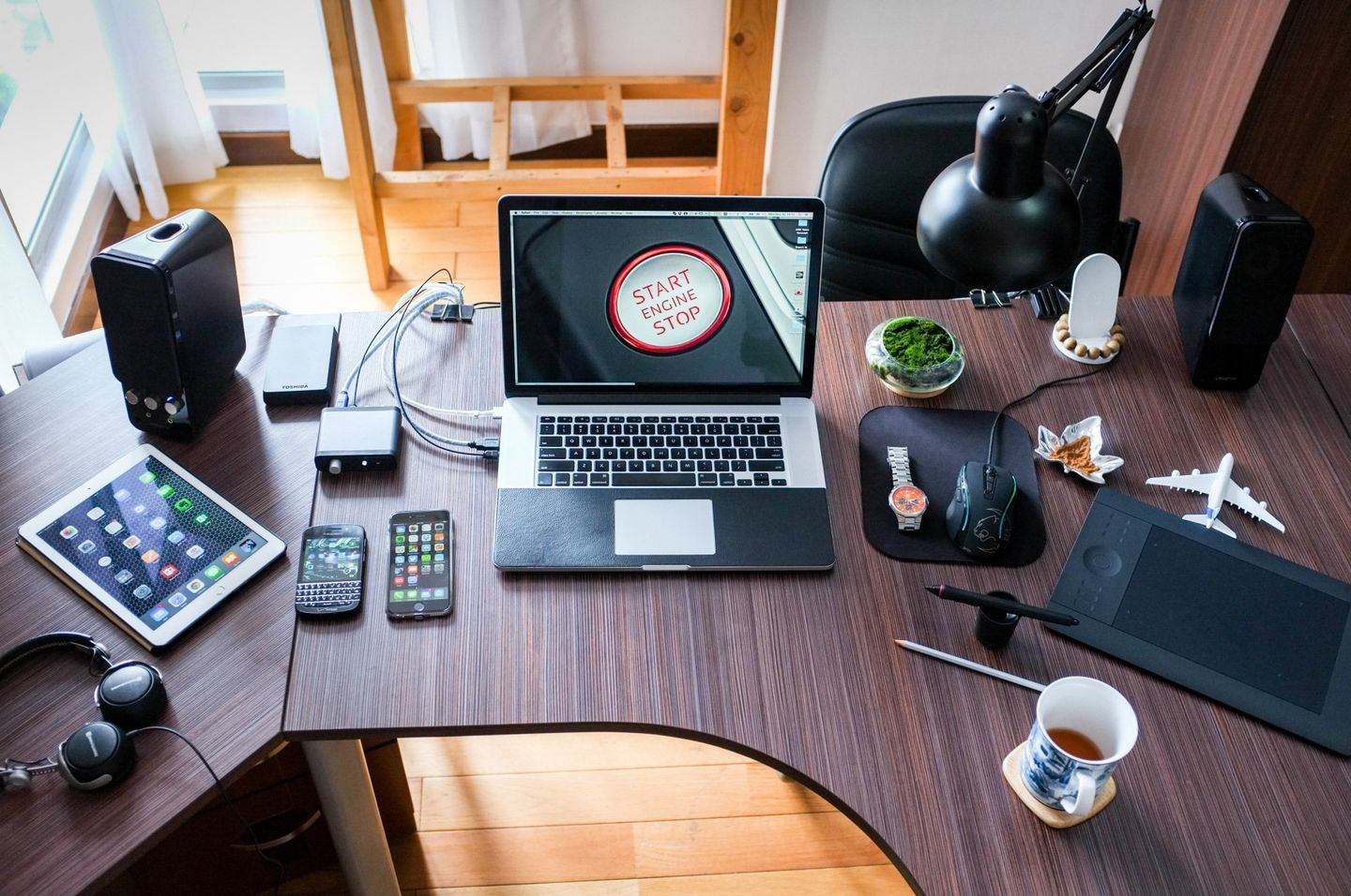 Laptop and gadgets on a wooden desk, including speakers, tablet, smartphones, headphones, and a drawing tablet.