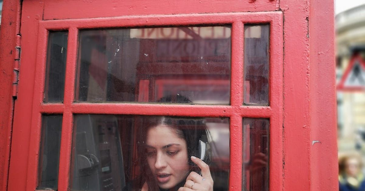 A woman is talking on a cell phone in a red telephone booth.
