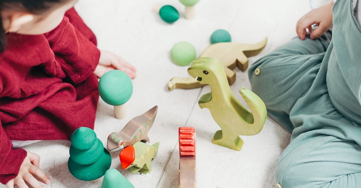 Two children playing with wooden dinosaur toys and trees on a white surface.