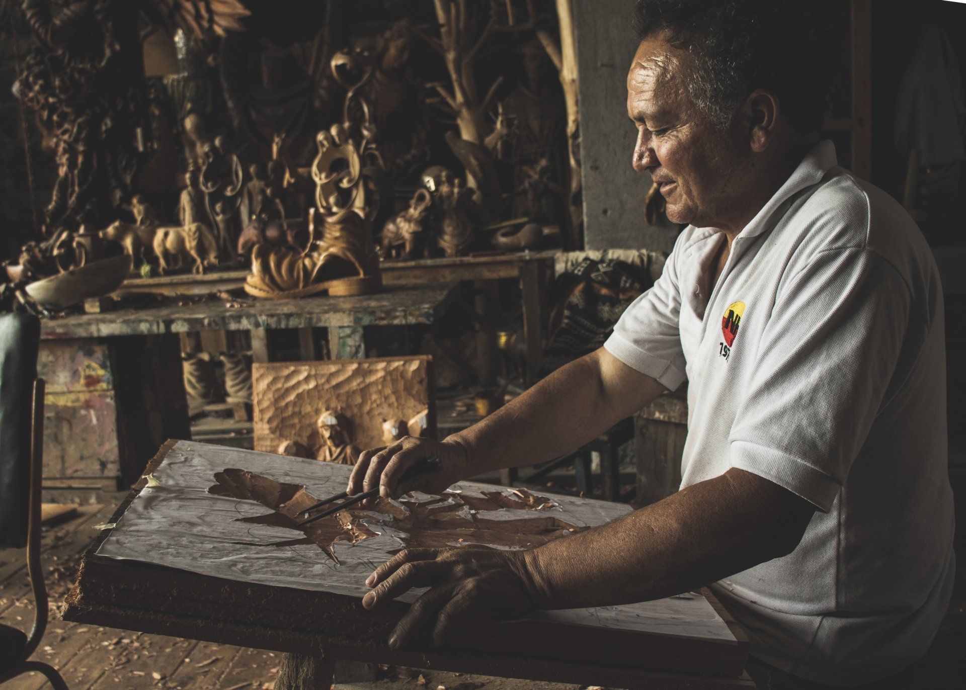 Man carving wood with tools at a workbench, workshop setting; sculptures in background.