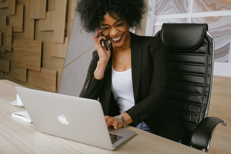 Woman with glasses, afro, blazer, talking on phone, smiling, working on a laptop at a desk.