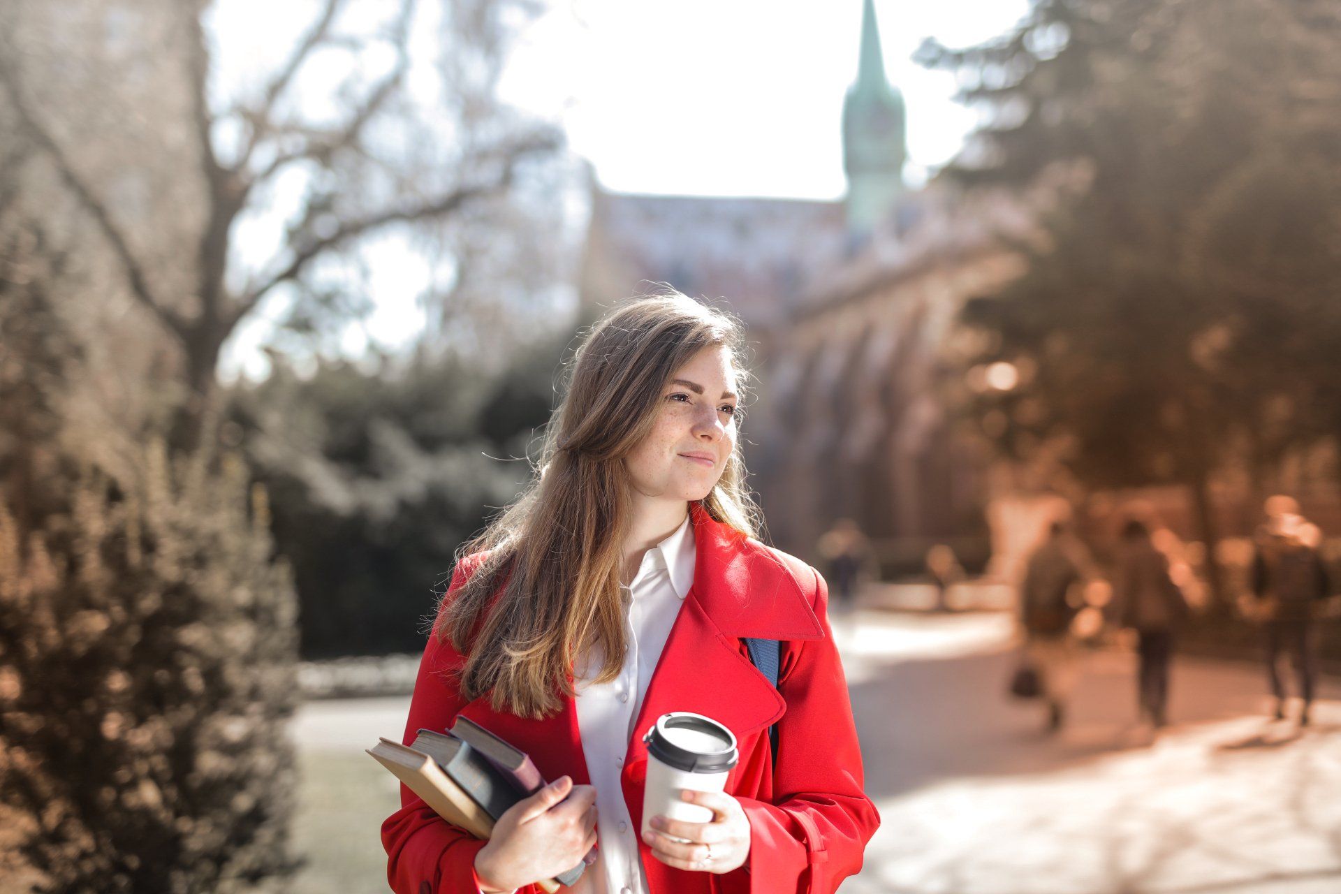 Teen girl in red blazer holding a cup of coffee and standing on a college campus looking around.