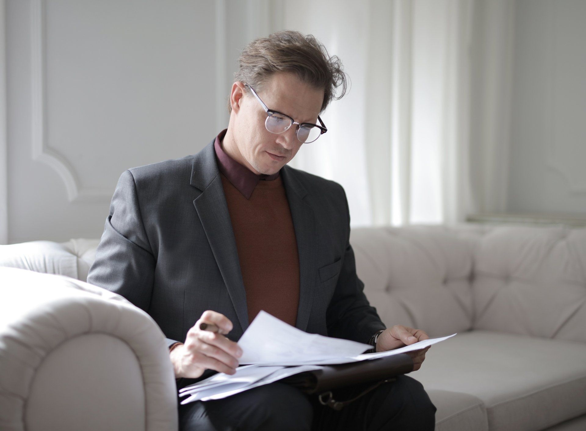 A man in a suit and glasses is sitting on a couch looking at papers.