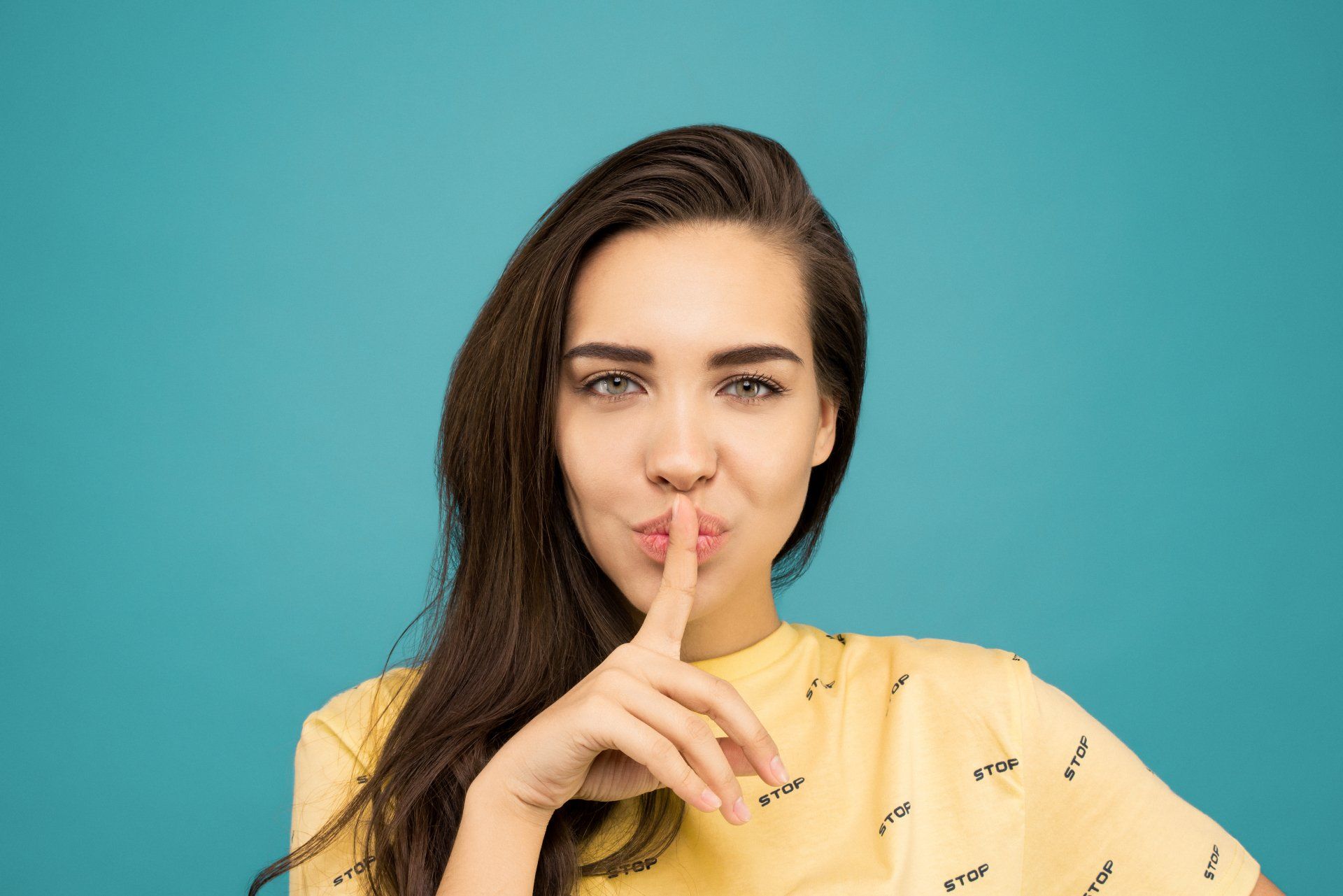 Een vrouw in een geel shirt houdt haar vinger tegen haar lippen.