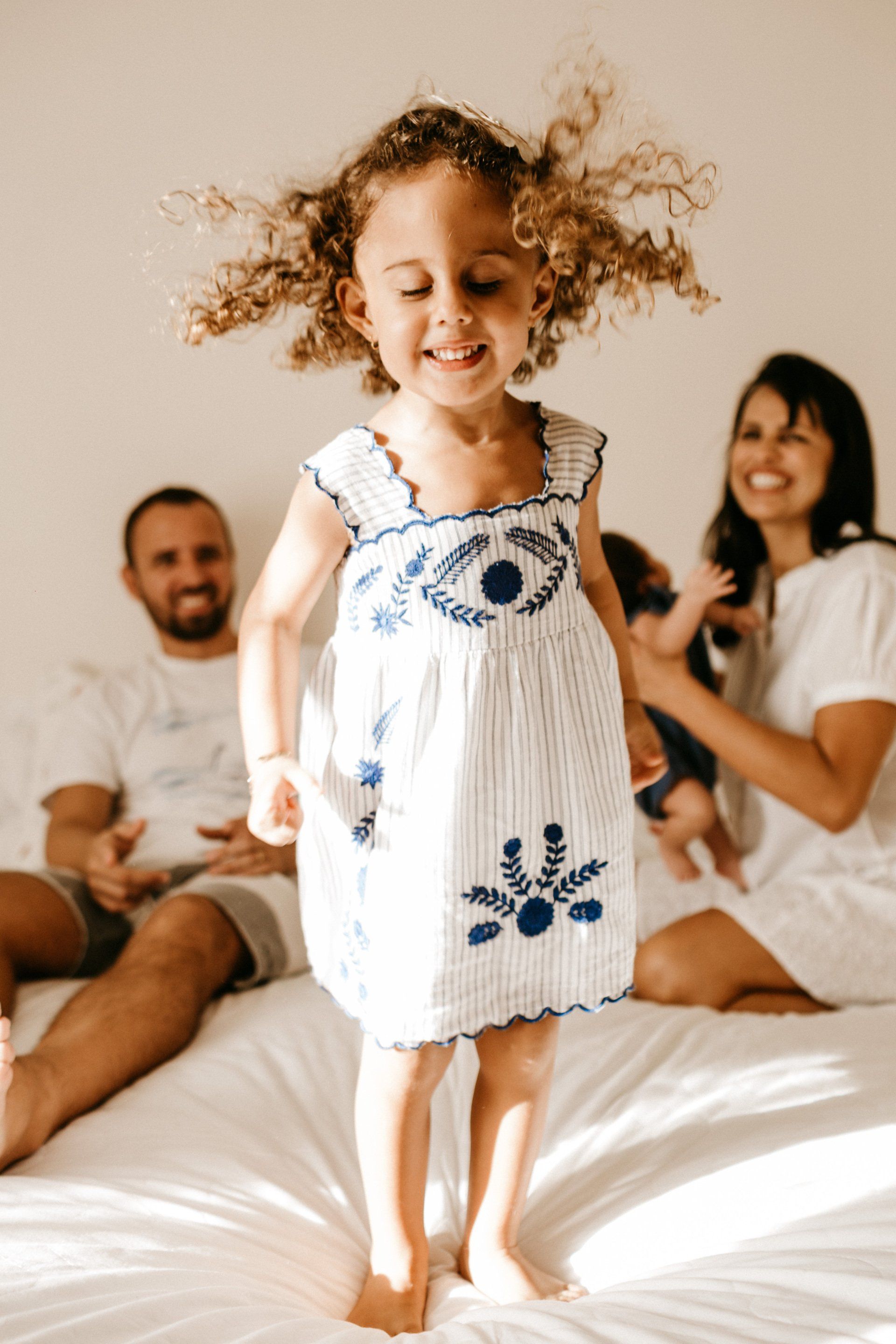 A little girl in a white dress is jumping on a bed.