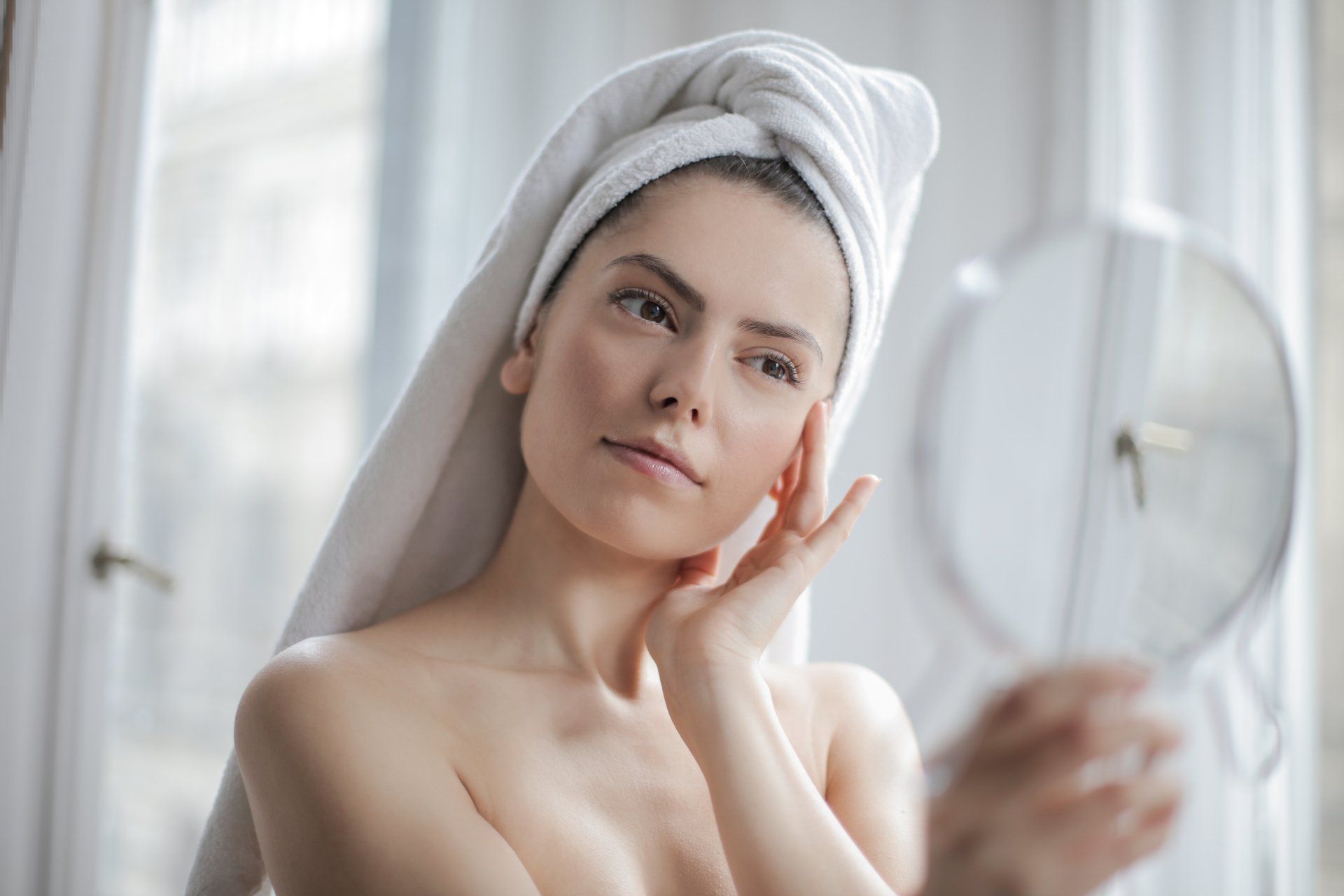 woman with white towel on head looks at her face with handheld mirror