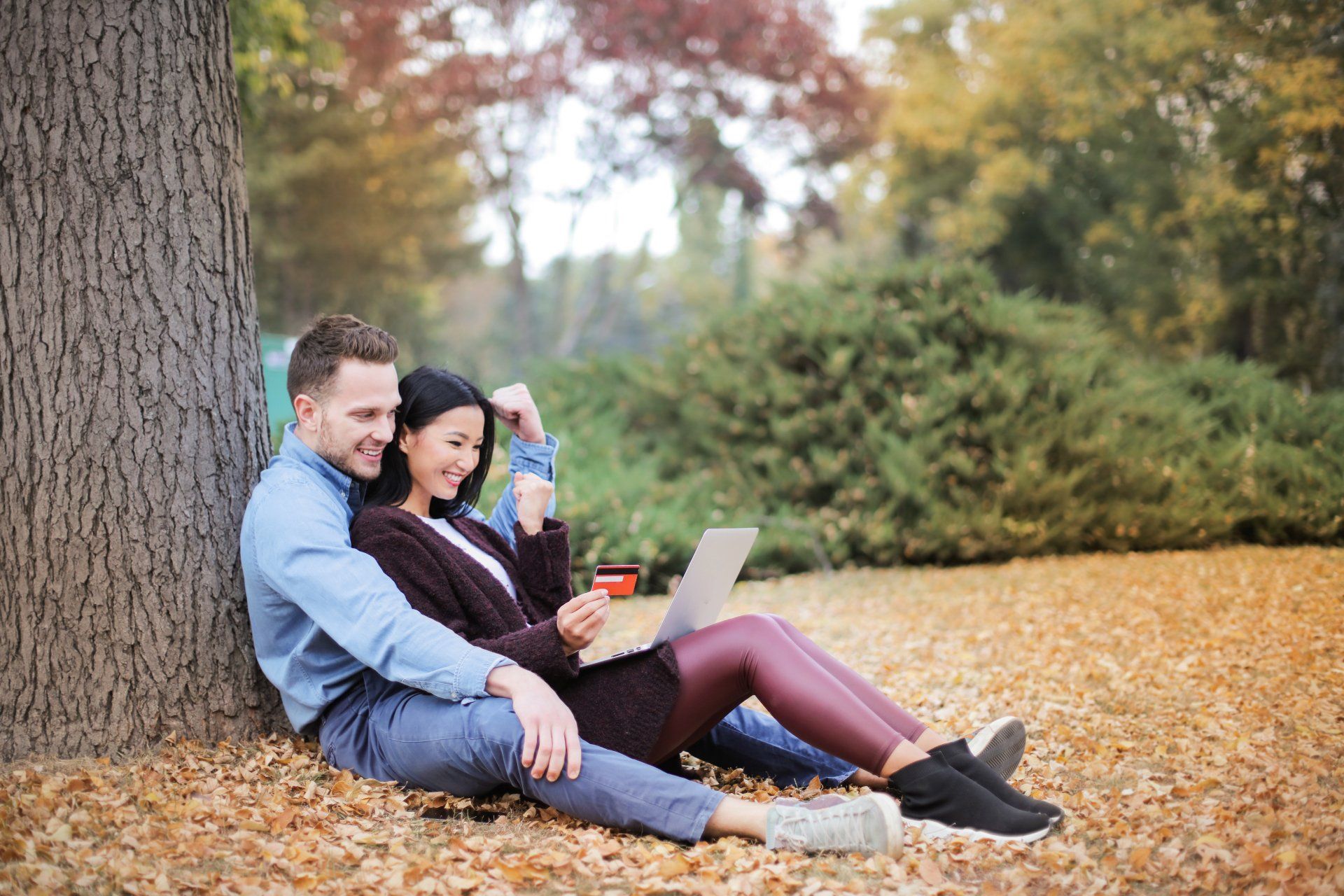 A man and a woman are sitting under a tree looking at a laptop.