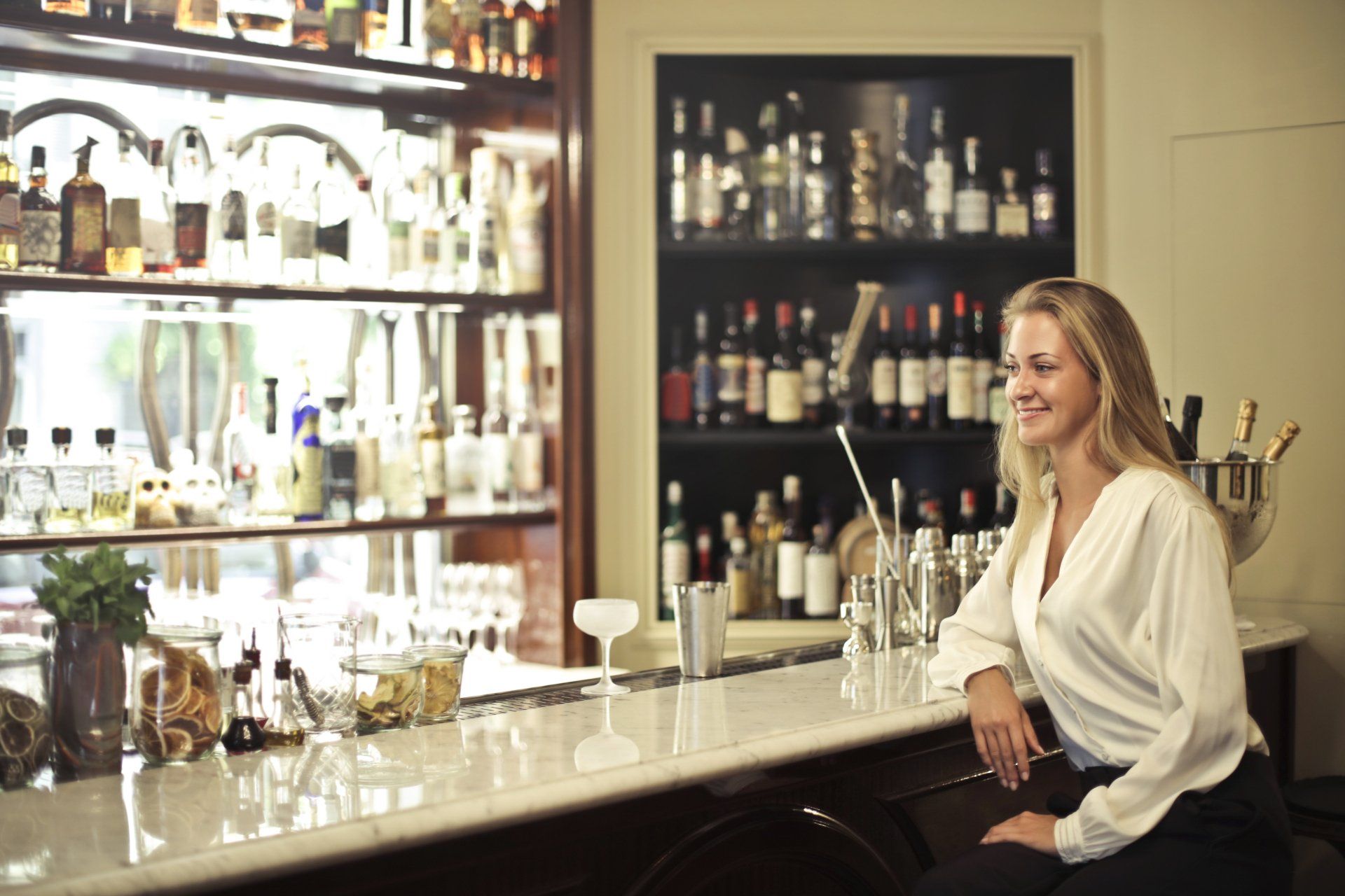 A woman is sitting at a bar looking at bottles of alcohol.