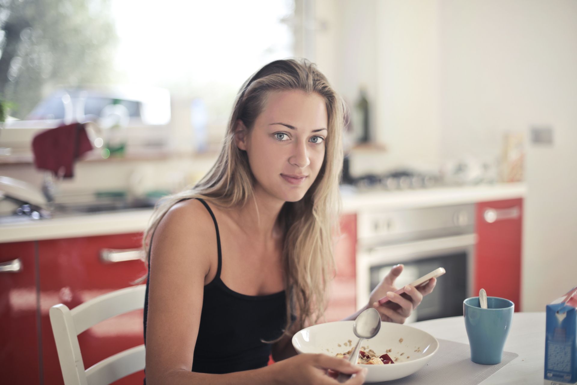 a woman is sitting at a table eating cereal and looking at her phone.