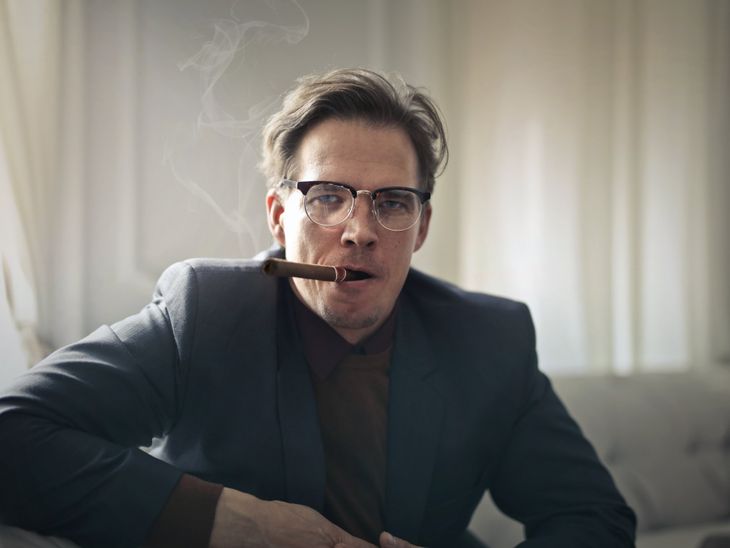 A middle aged white man in a suit , sitting on a couch, with a cigar in his mouth