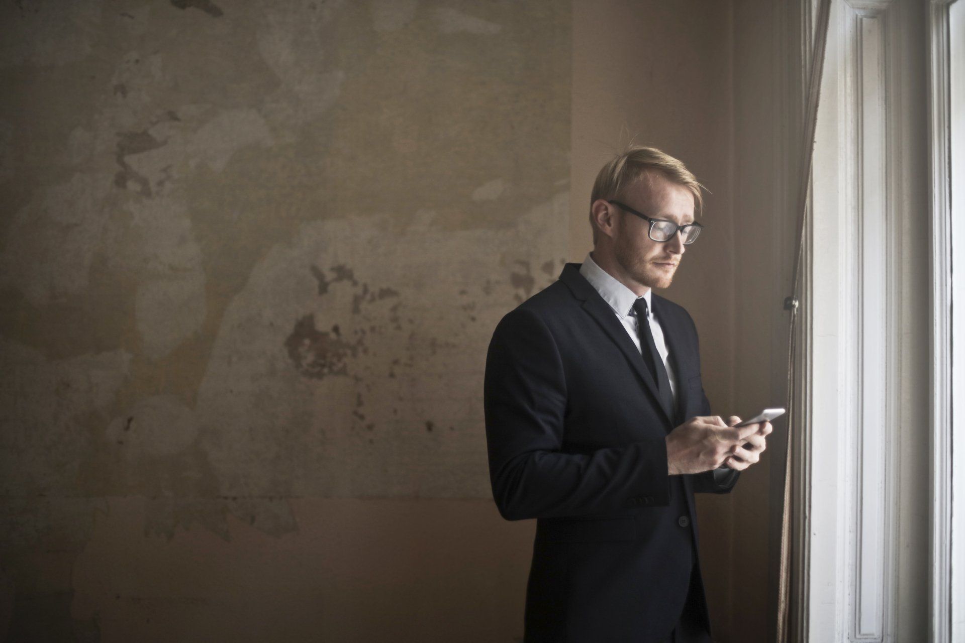 Man in suit, glasses, looking at phone near a window with curtains; brown wall.
