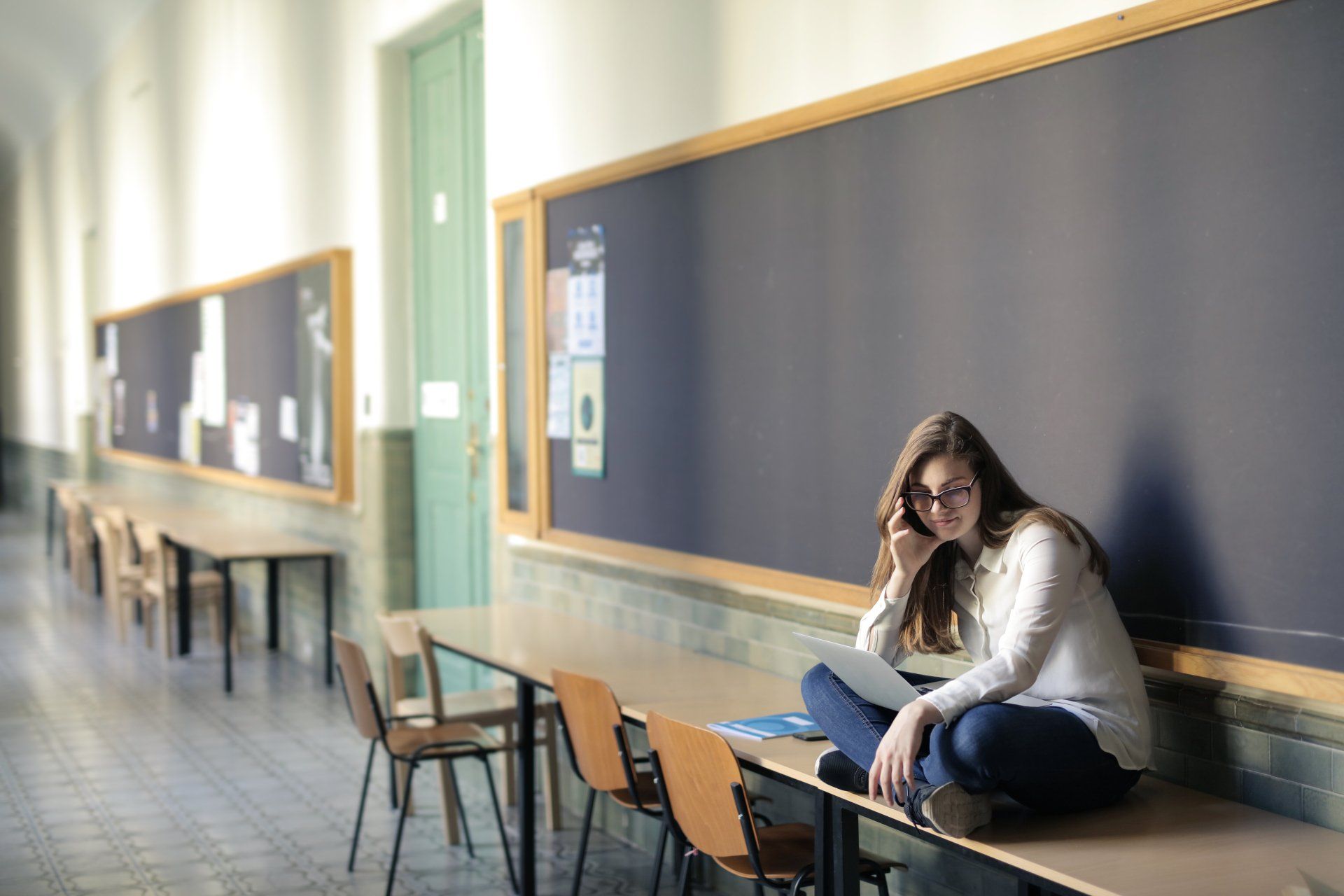 A woman is sitting on a desk in a classroom talking on a cell phone.