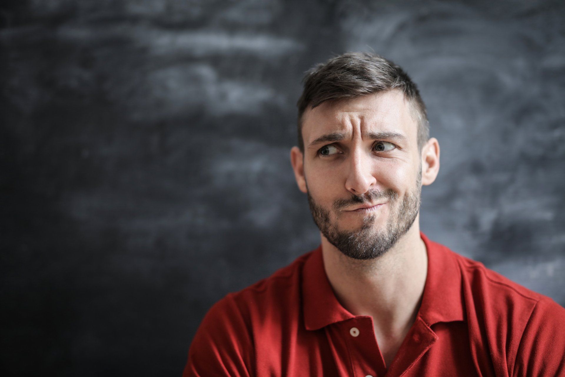 A man in a red shirt is making a funny face in front of a blackboard.