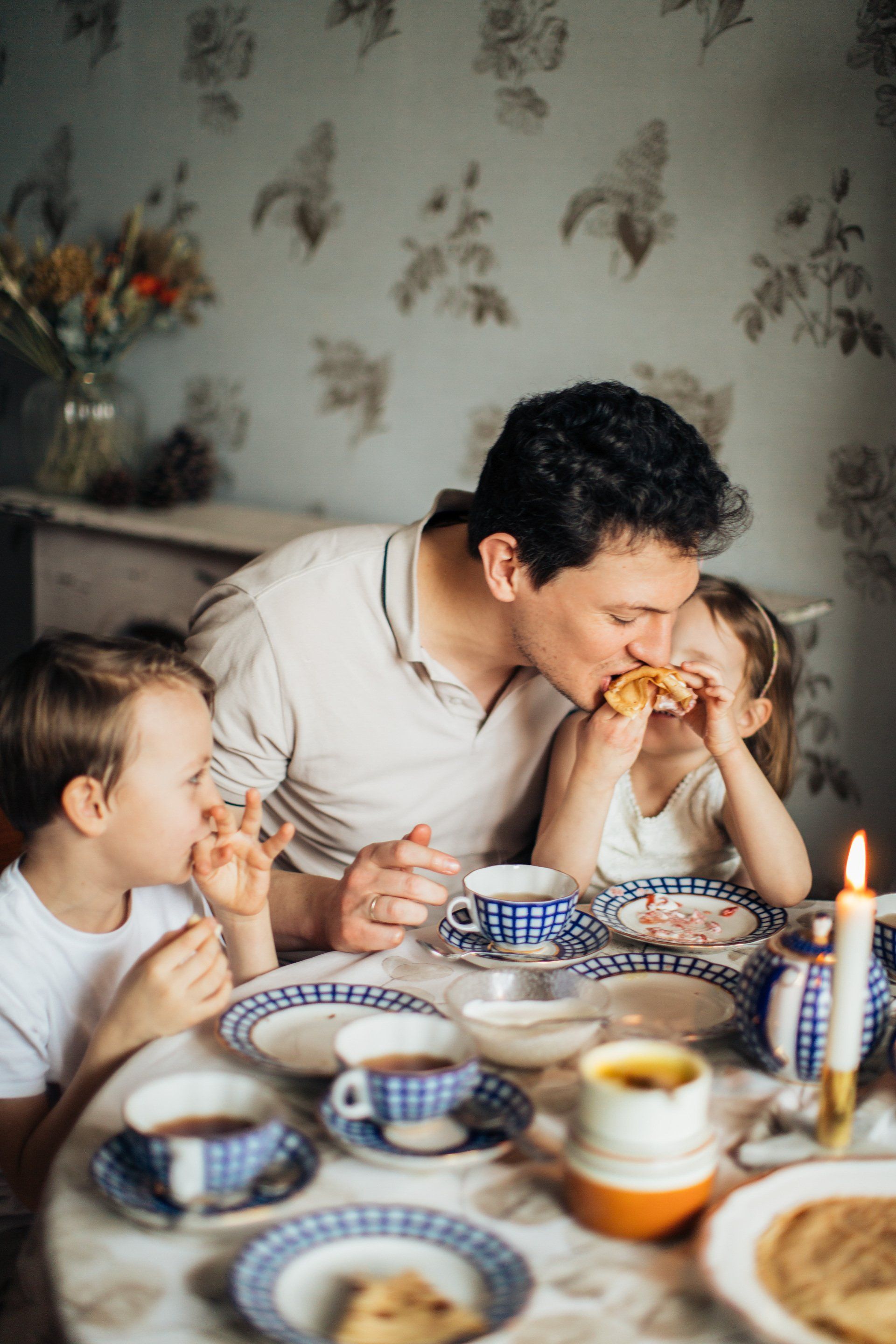 Father kissing child eating food at a table with another child; tea and plates on the table.