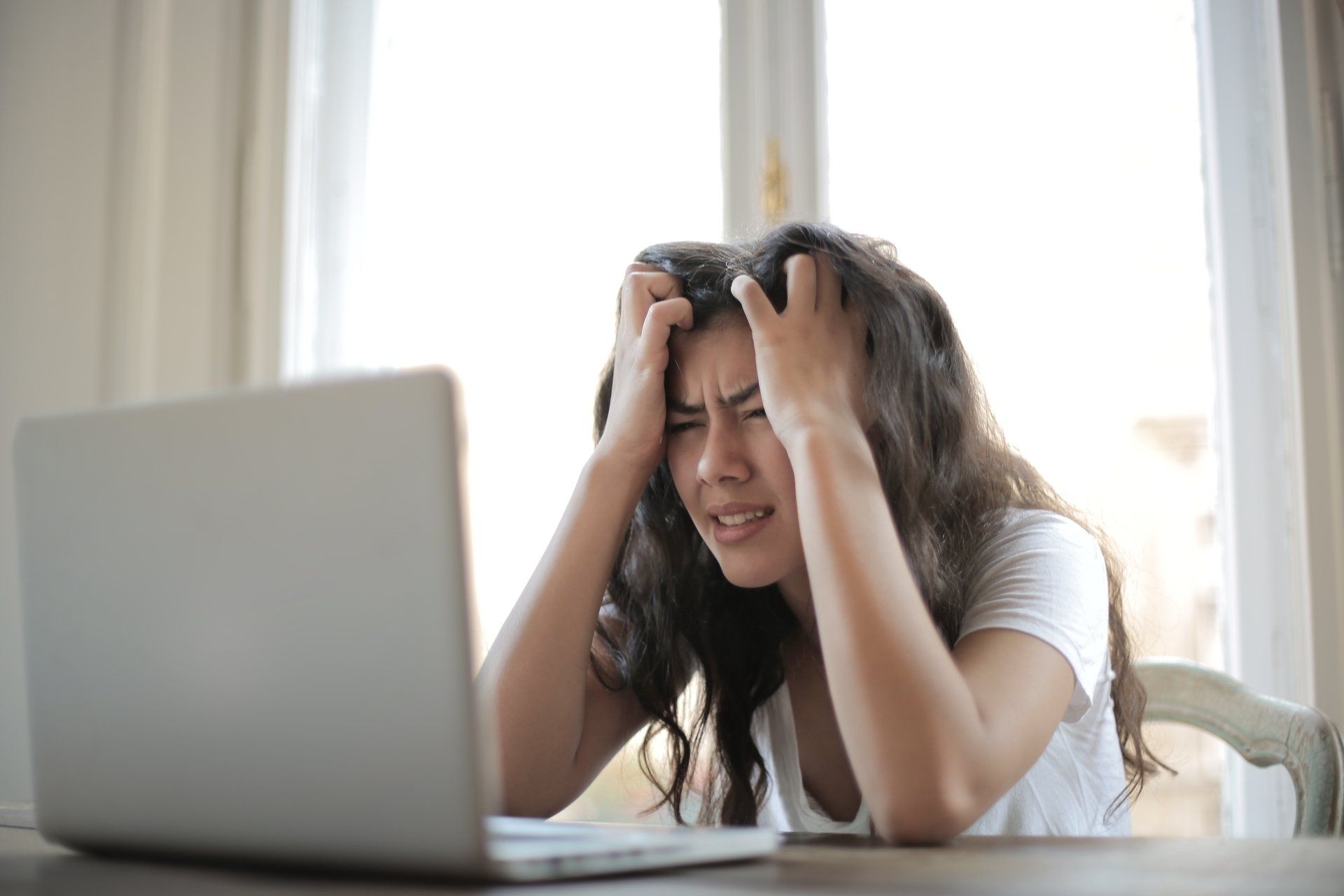 A woman is sitting at a table with her hands on her head in front of a laptop computer.