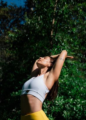 A woman in a white crop top and yellow pants is doing yoga in the woods.