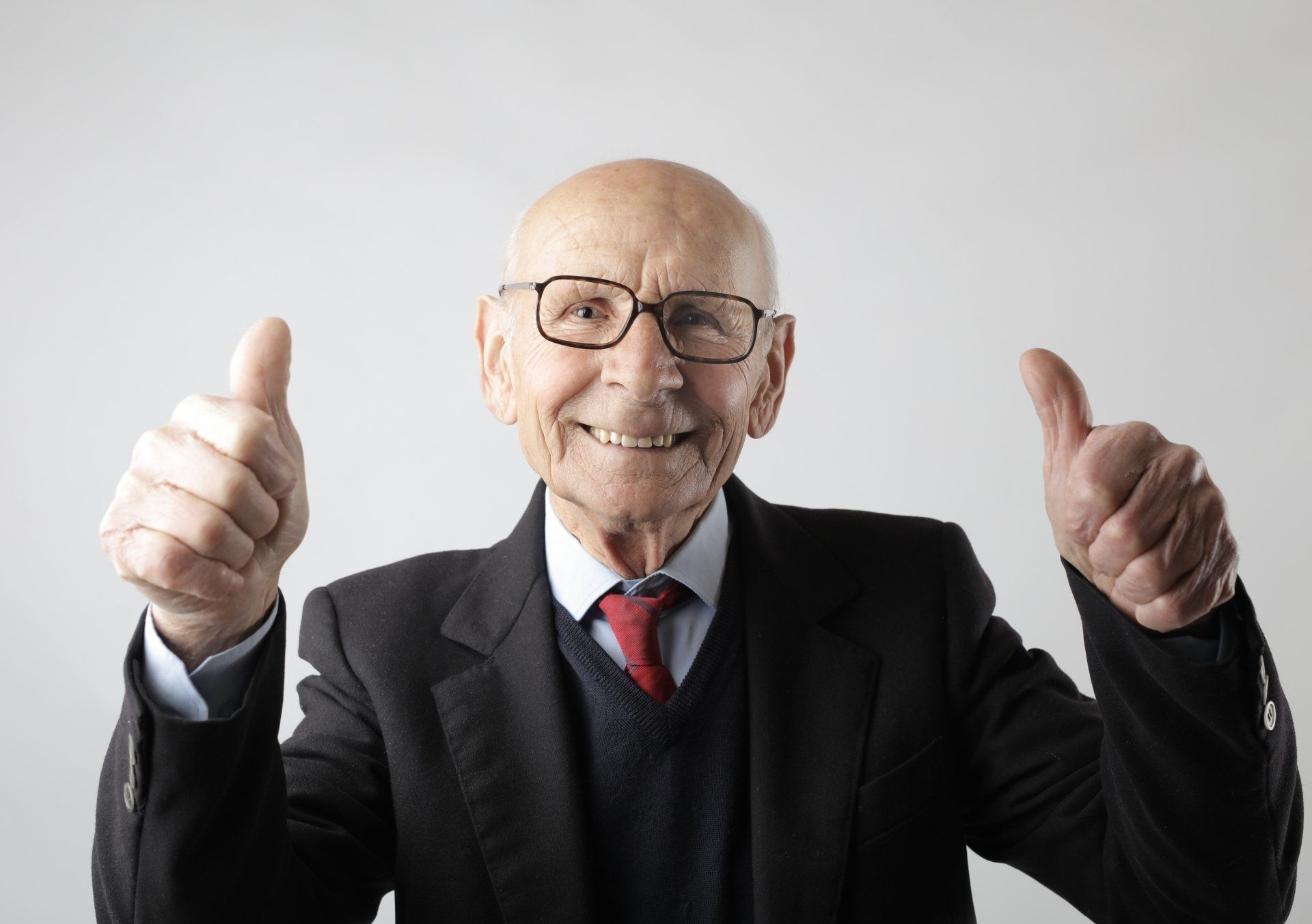 An elderly man in a suit and tie is giving two thumbs up.
