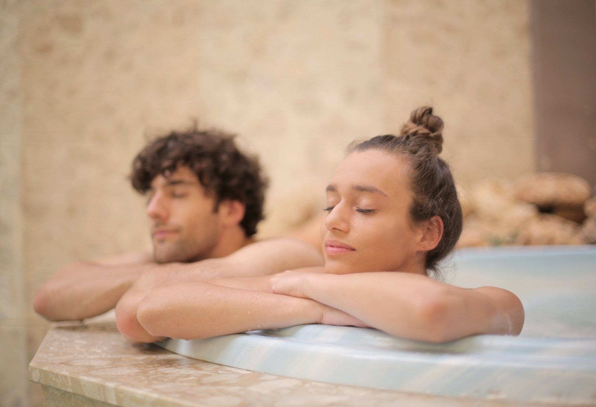 Man and woman relaxing in a spa. Both have their eyes closed, leaning on the edge of a light-colored tub.