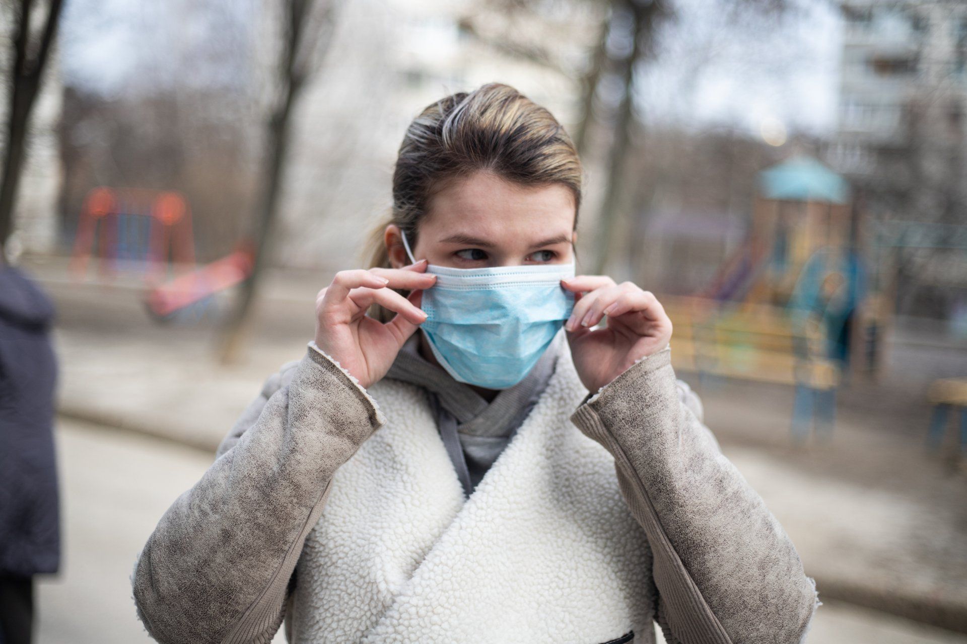 Woman adjusts blue face mask outdoors. Neutral coat, park background.