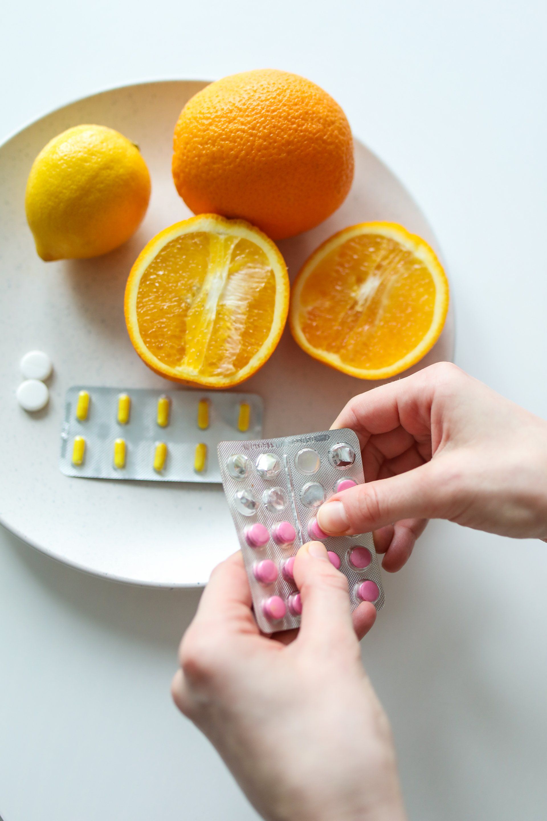 A person is holding a blister pack of pills in front of a plate of oranges and lemons.