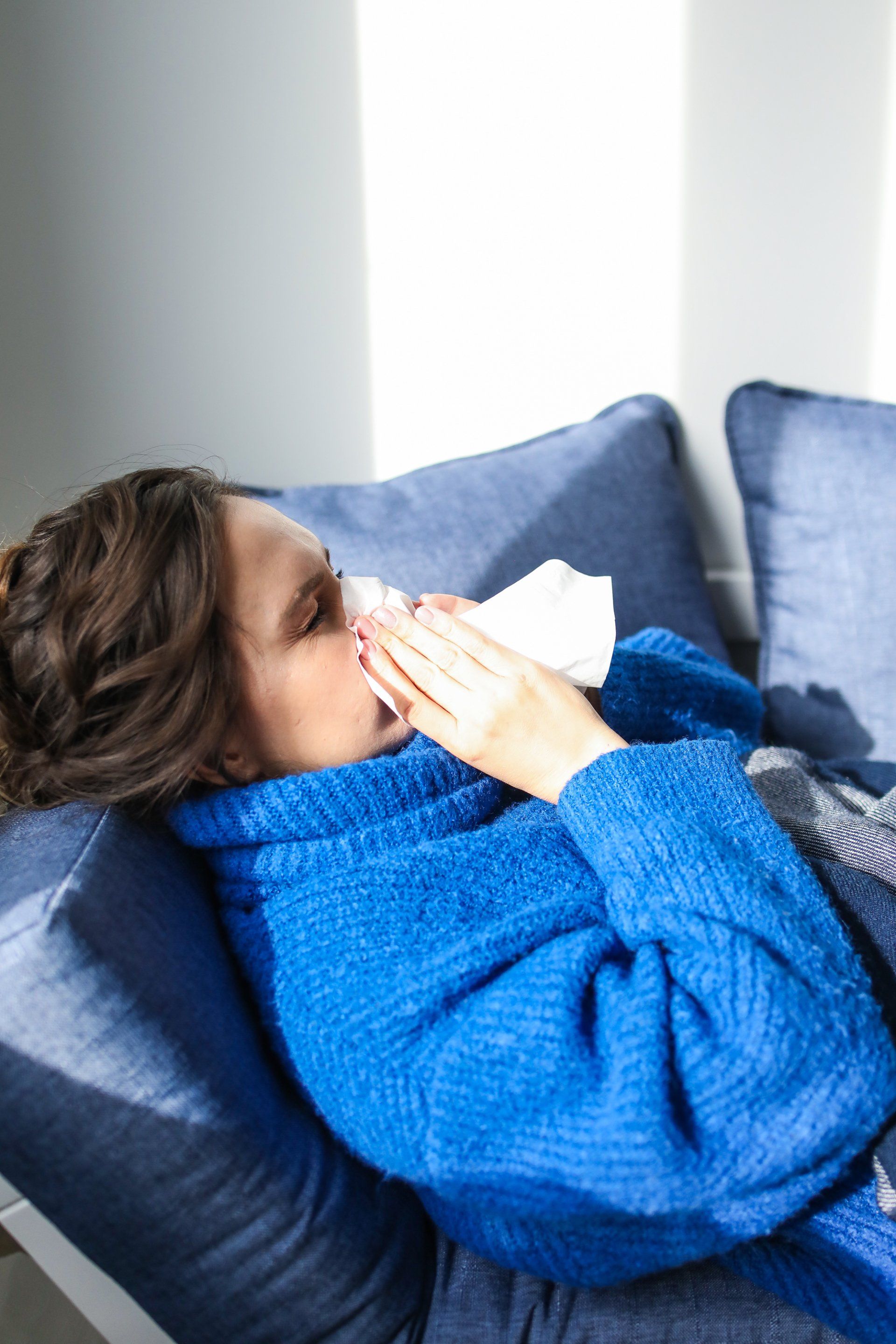 woman in blue robe laying on a couch sneezing into a napkin