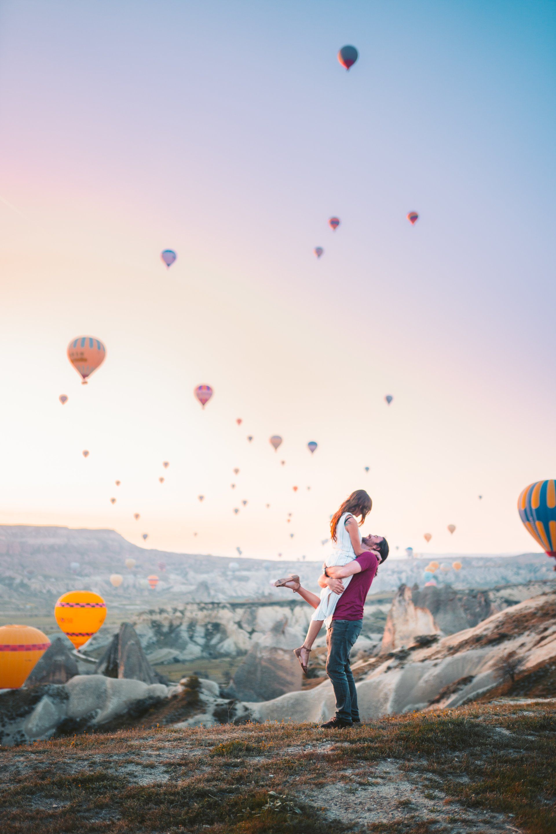A man is holding a woman in his arms in front of hot air balloons flying in the sky.