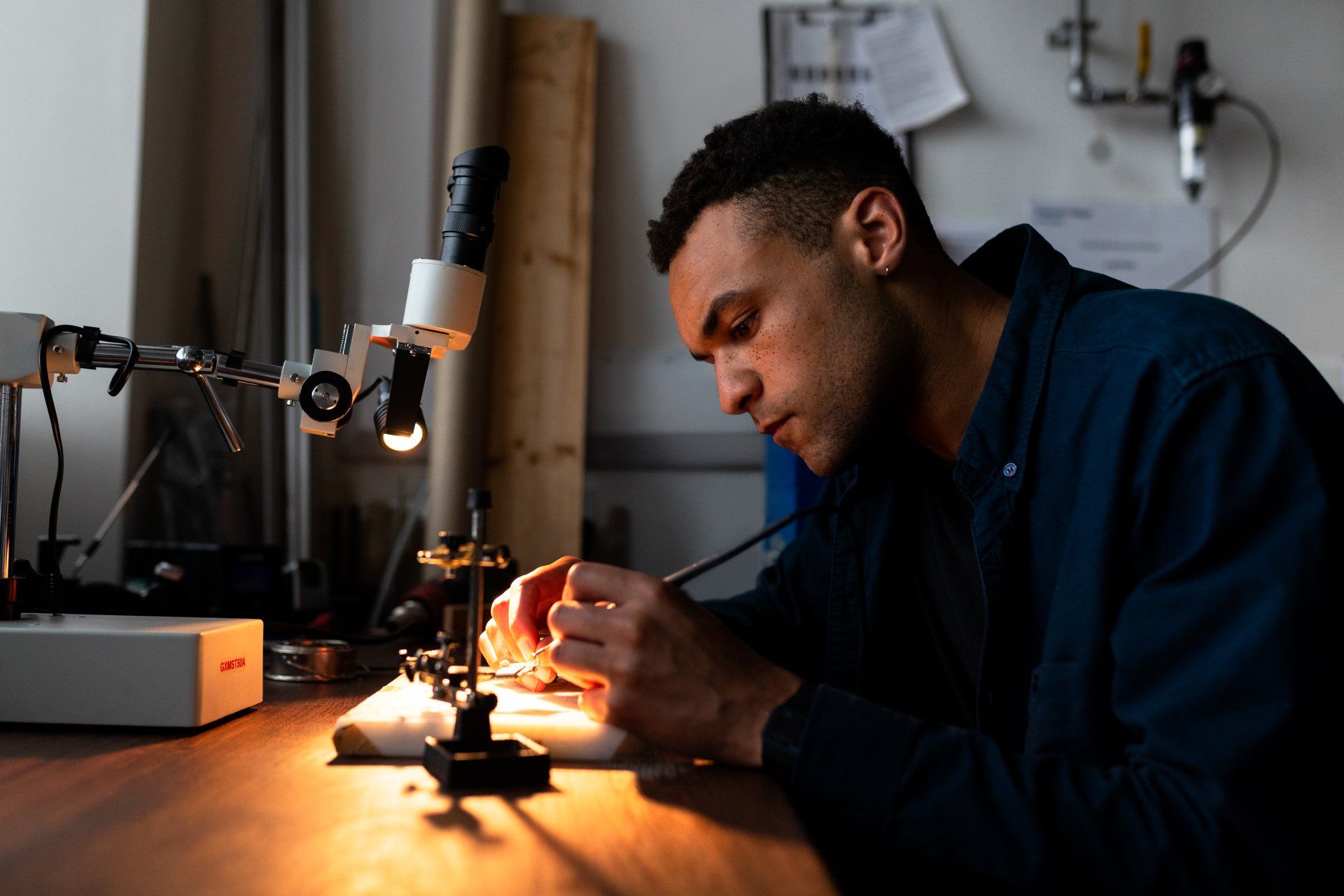 A person in a blue shirt works with a small tool under a magnifying lamp in a dimly lit workshop.
