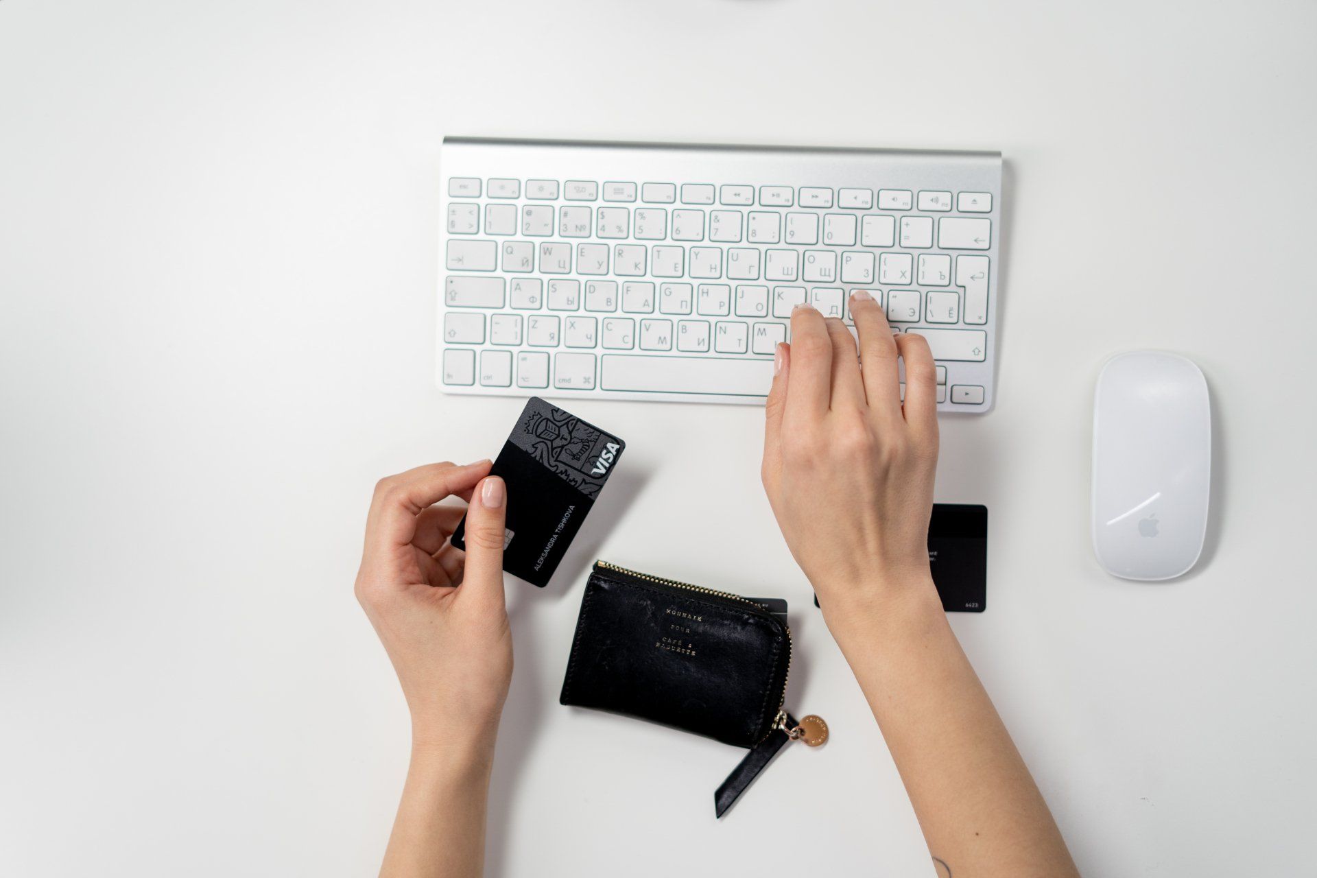 A keyboard, mouse and wallet on a table top while a person holding a credit card online shops.