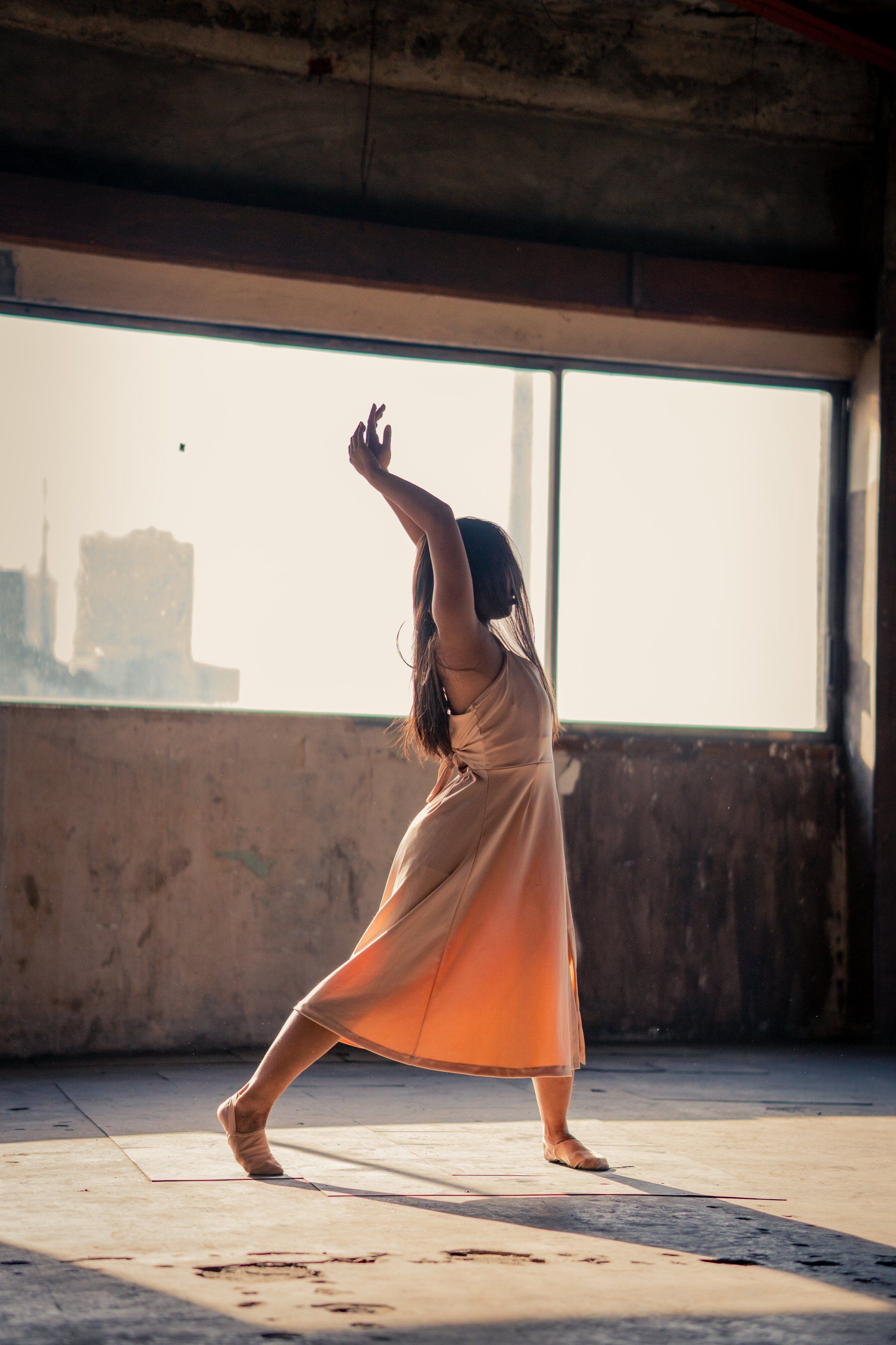 A woman in a dress is dancing in a room in front of a window.