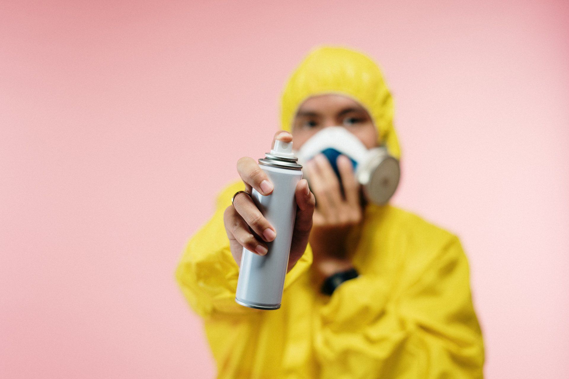 man wearing yellow protection suit in pink background wearing a mask and holding pest spray