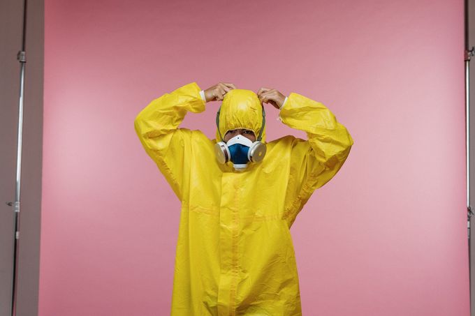 A man in a yellow protective suit and mask is standing in front of a pink background.