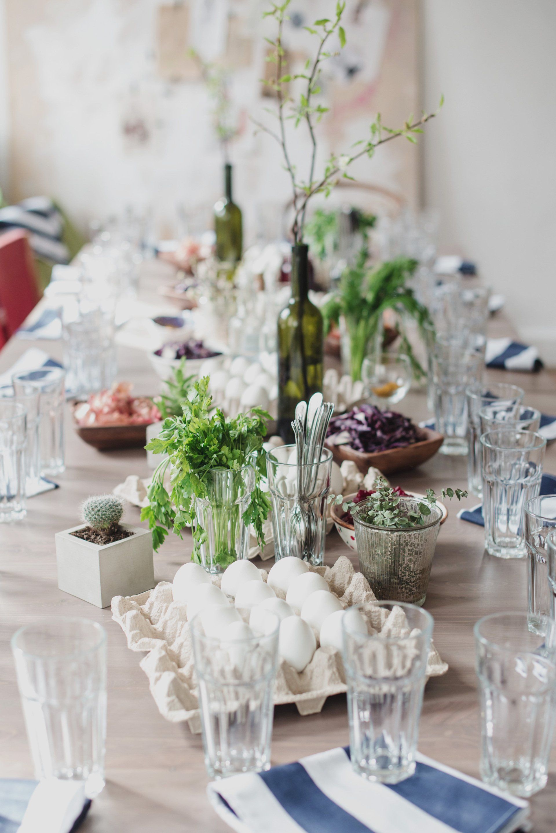 A long table set for a dinner party with glasses , bowls , plants and a bottle of wine.