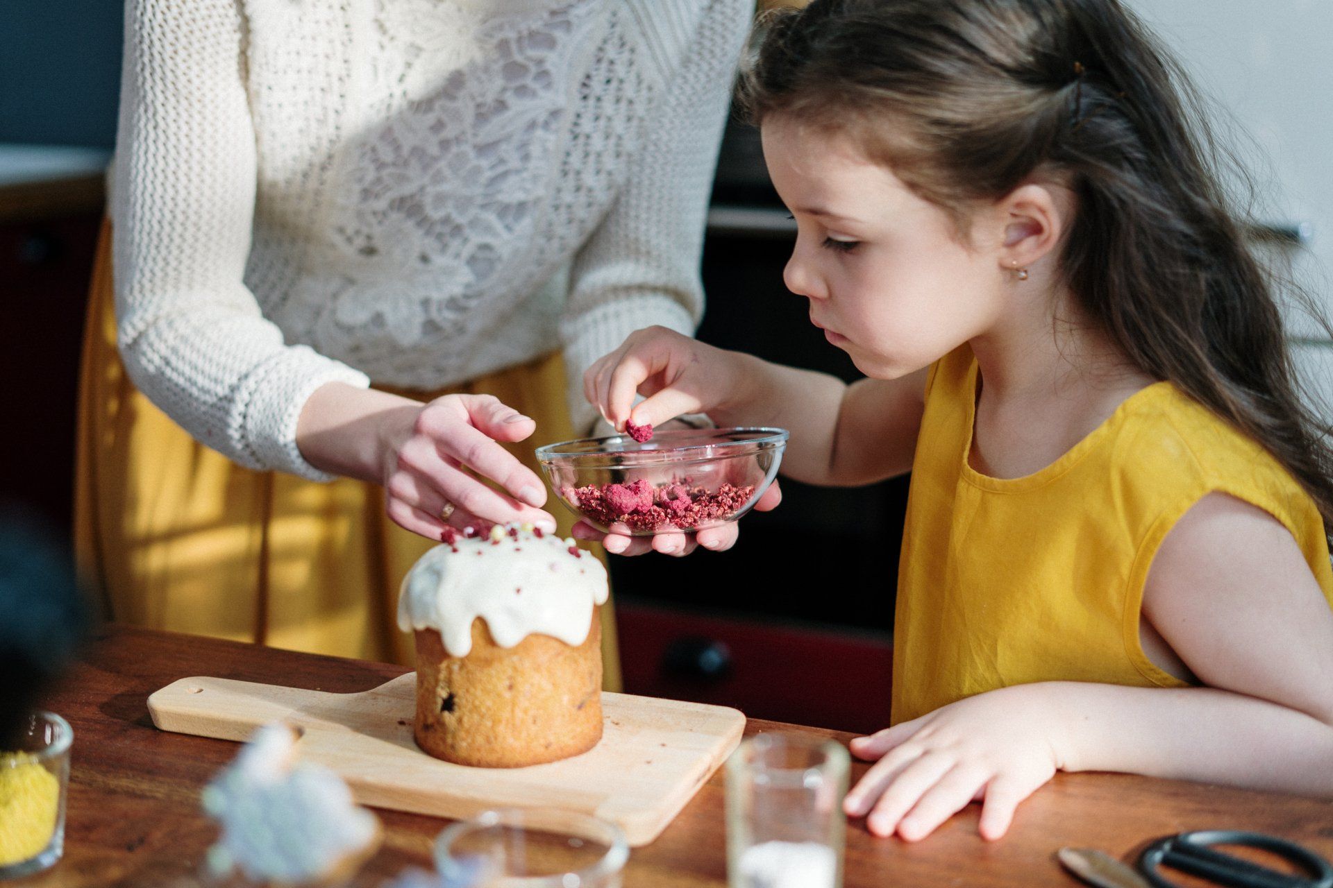 A woman and child decorate a frosted cake with sprinkles in a kitchen. The child carefully sprinkles.