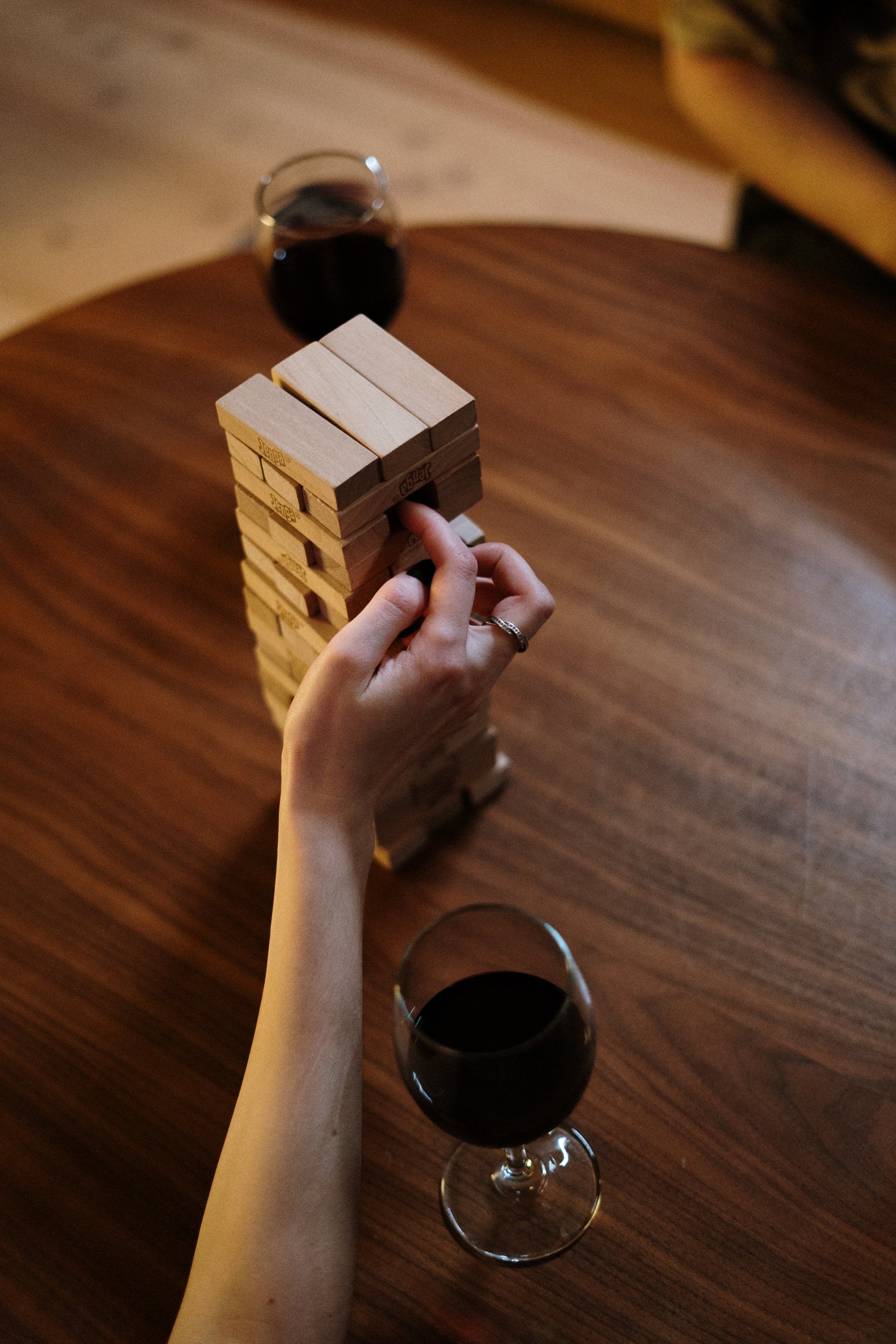 A game of Jenga on a table with someones hand pushing a brick out. A glass of red wine at hand.