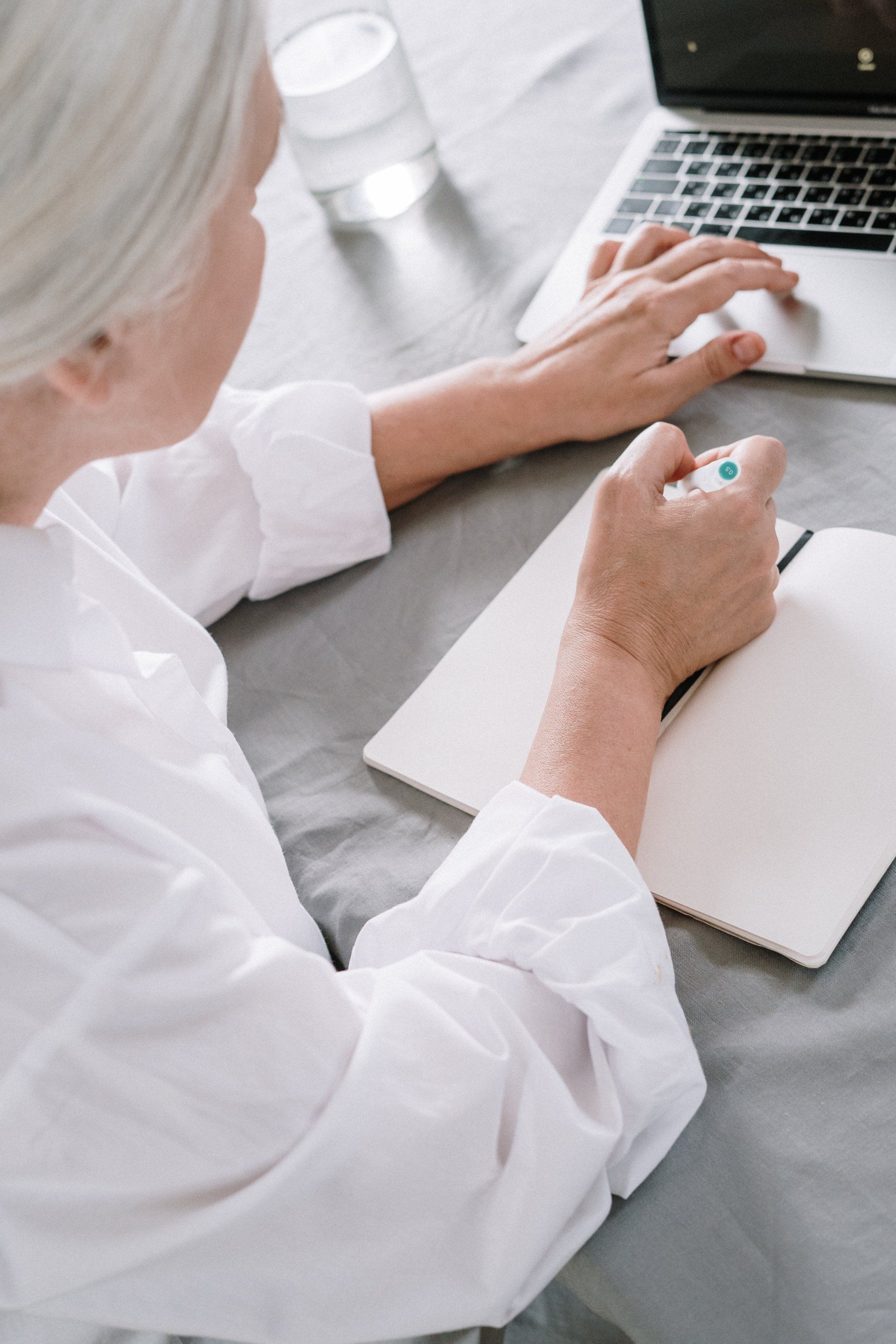 A woman is sitting at a table using a laptop computer and writing in a notebook.