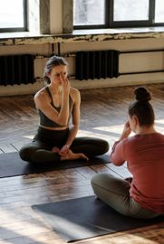 Two women are sitting on yoga mats on the floor in a room.