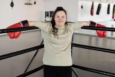 A woman with down syndrome is standing in a boxing ring with her arms outstretched.