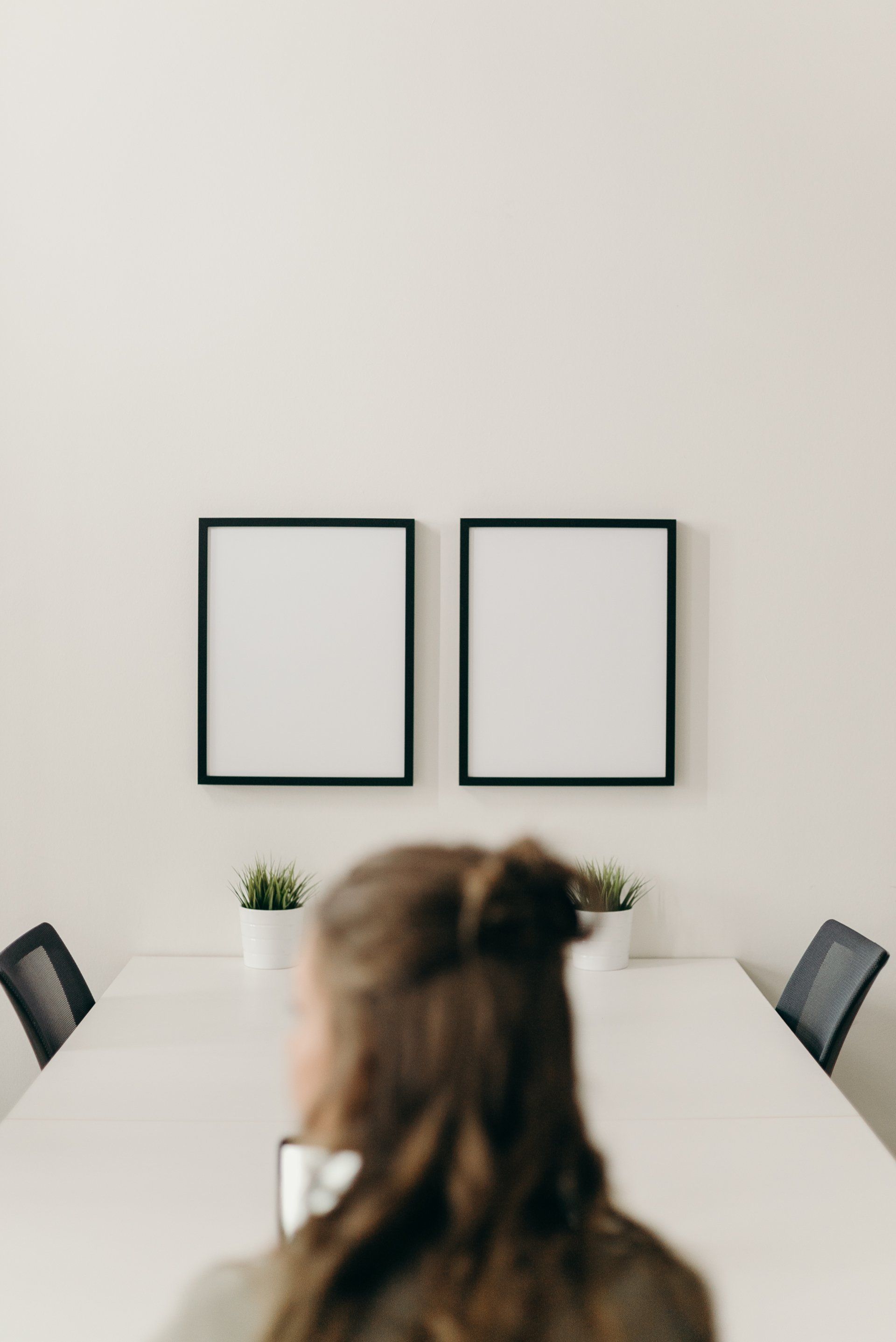 Woman facing a white table with two blank framed prints and plants, in an office setting.