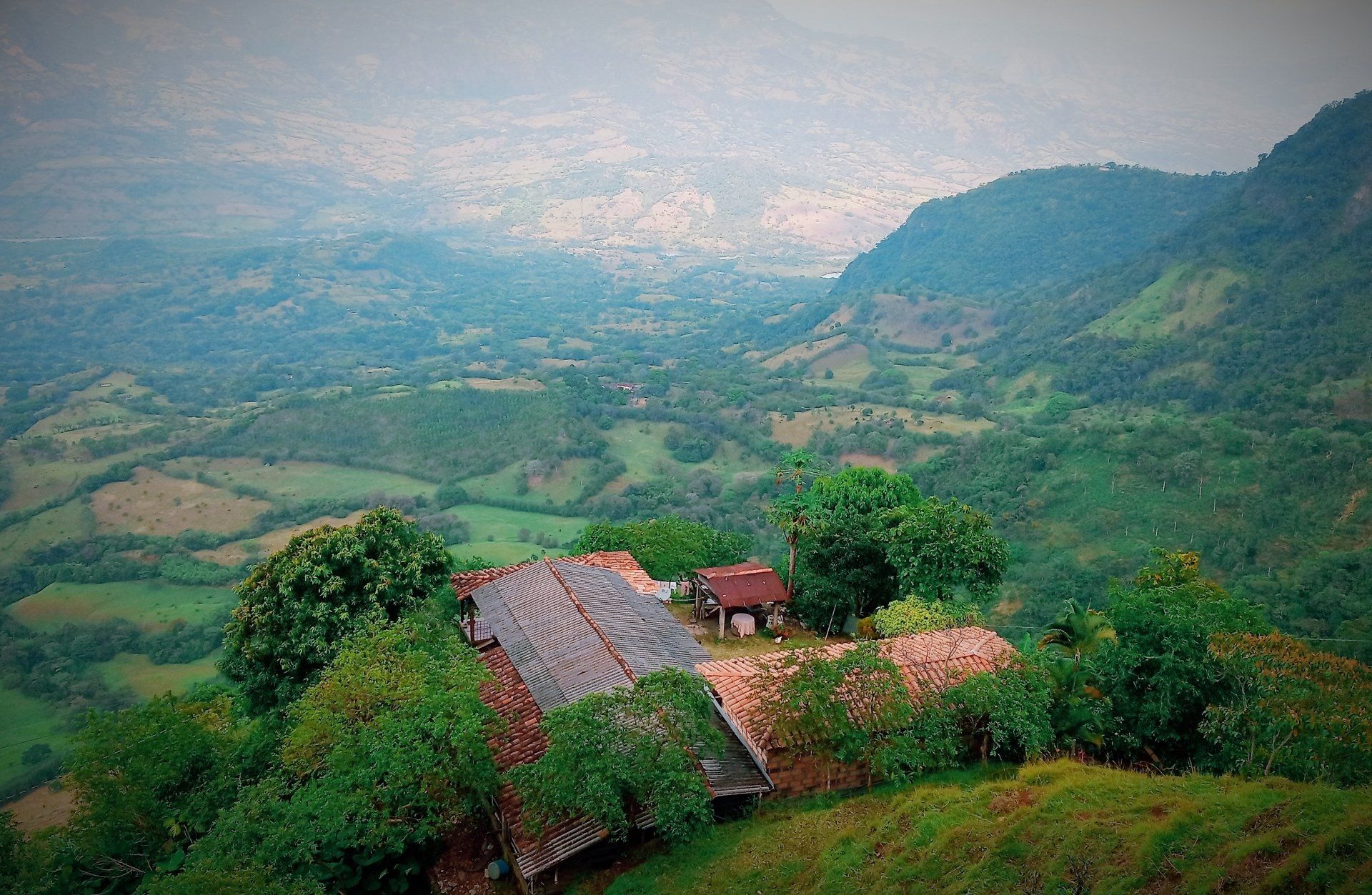 Overlooking valley, hillside buildings with red tile roofs. Lush green landscape under cloudy sky.