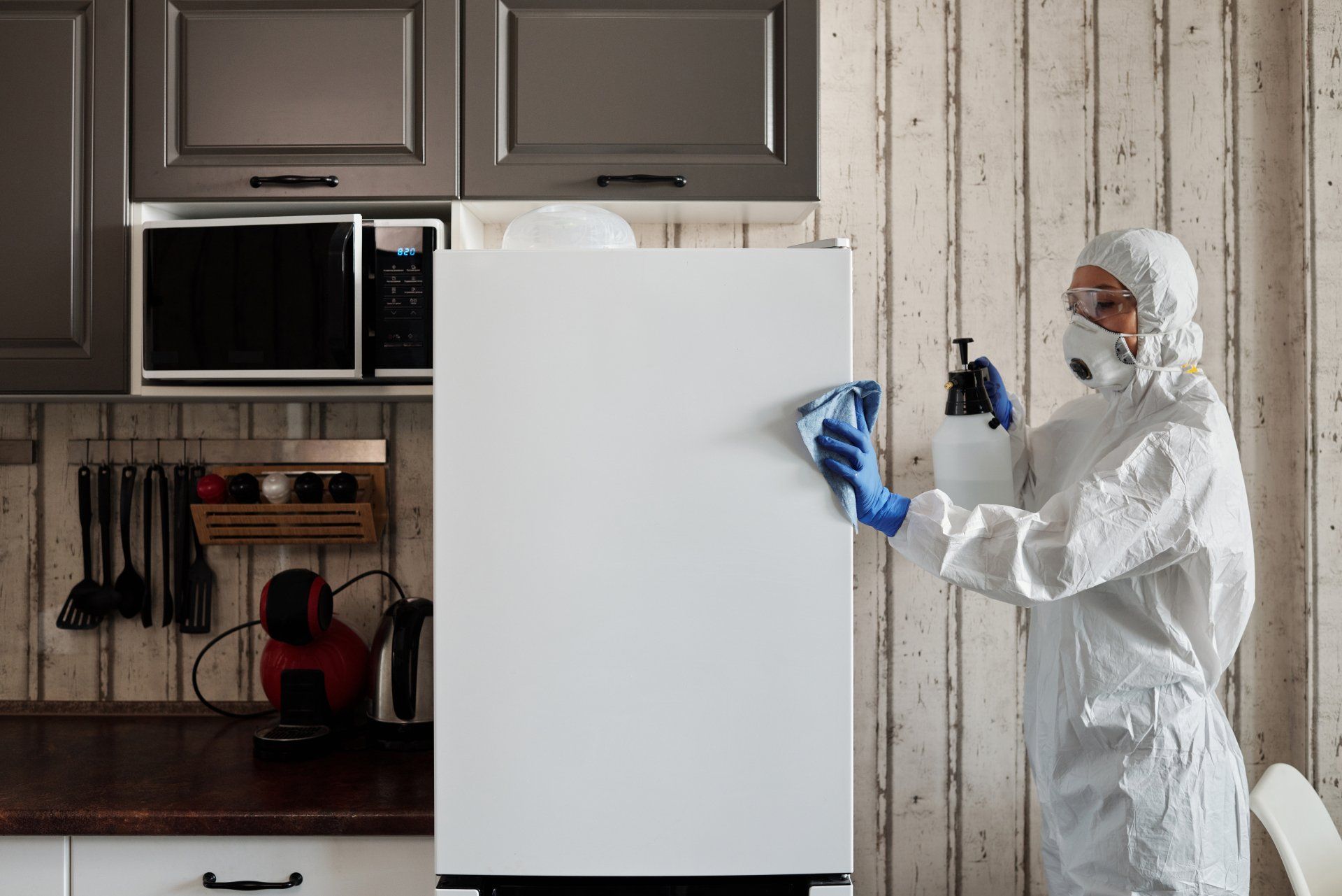 A person in a protective suit is cleaning a refrigerator in a kitchen.