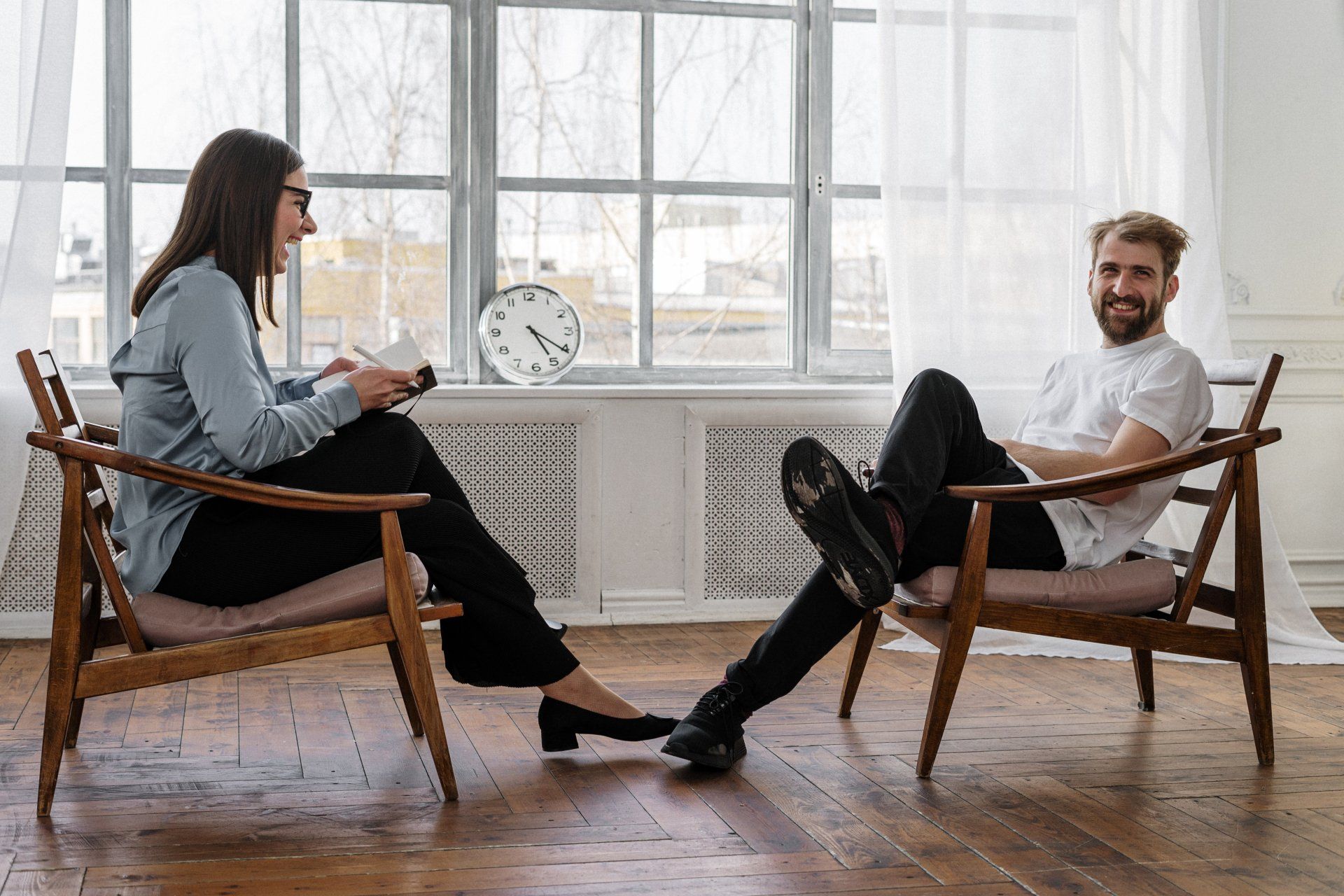 a man and a woman sit in chairs in front of a window with a clock talking about integrative therapy