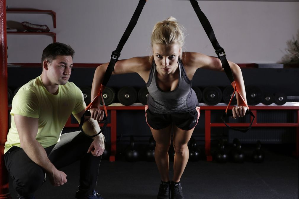 A man is watching a woman do trx exercises in a gym.