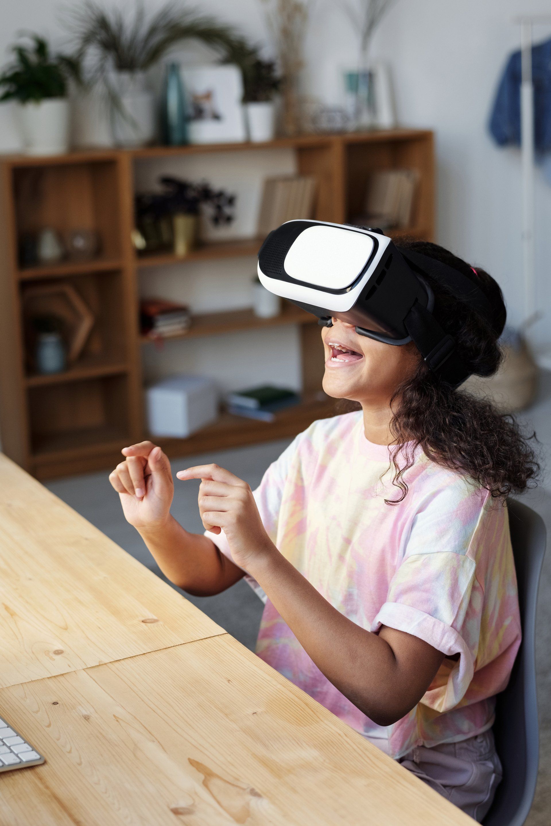 Girl wearing VR headset, sitting at desk, gesturing with hands, smiling. Wooden shelves in background.
