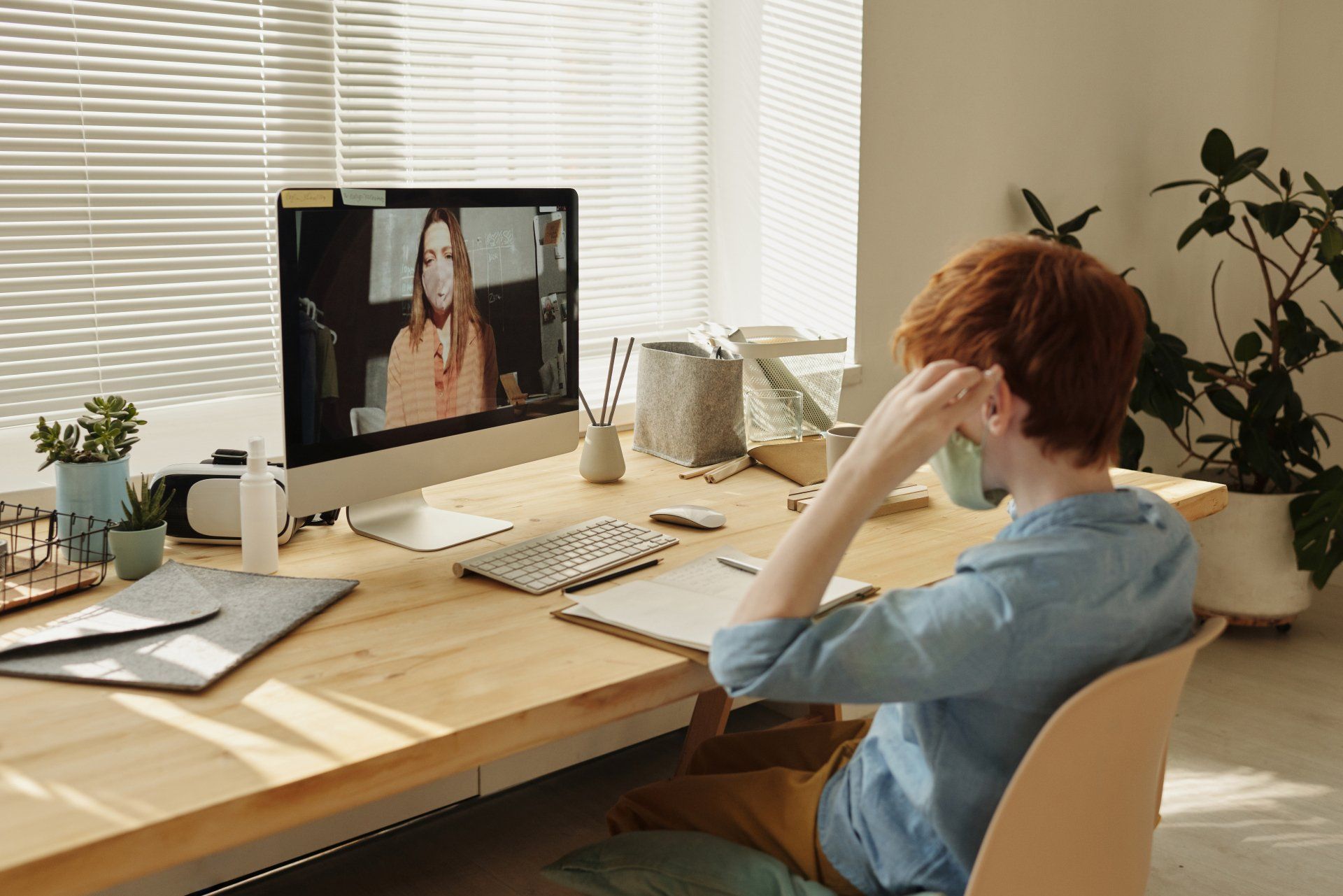 Une personne aux cheveux roux portant une chemise bleue regarde une femme sur un écran d'ordinateur à un bureau.