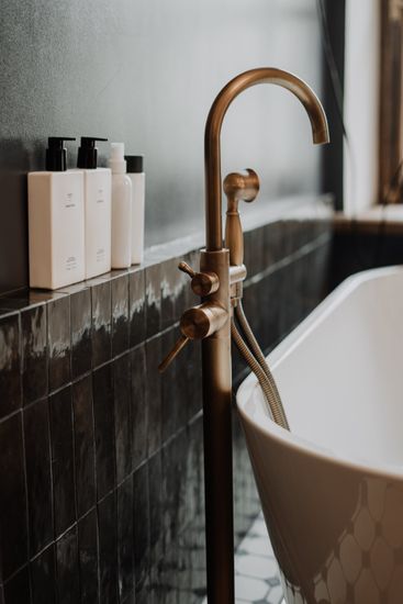 Solo standing tub with gold faucet standing in front of a black tile accent wall