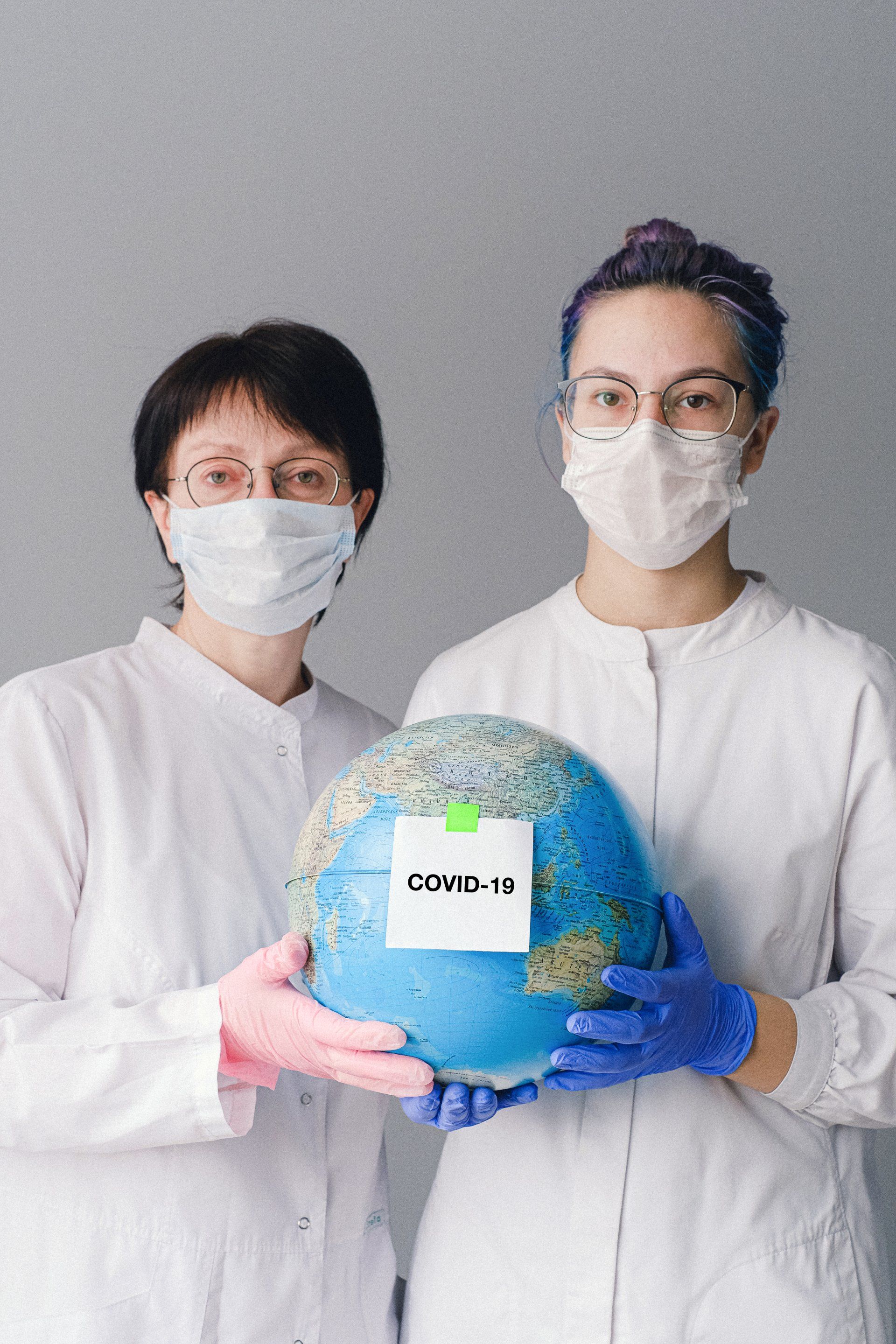 Two female doctors wearing masks and gloves are holding a globe.