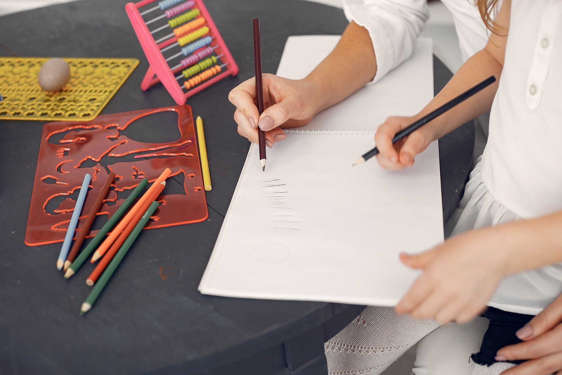 A woman and a child are sitting at a table drawing with pencils.