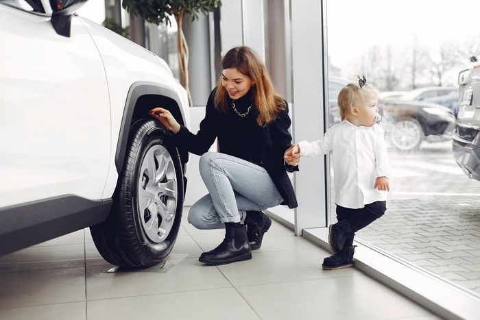 Woman kneels by a white car, holding a child’s hand in a bright showroom or driveway.