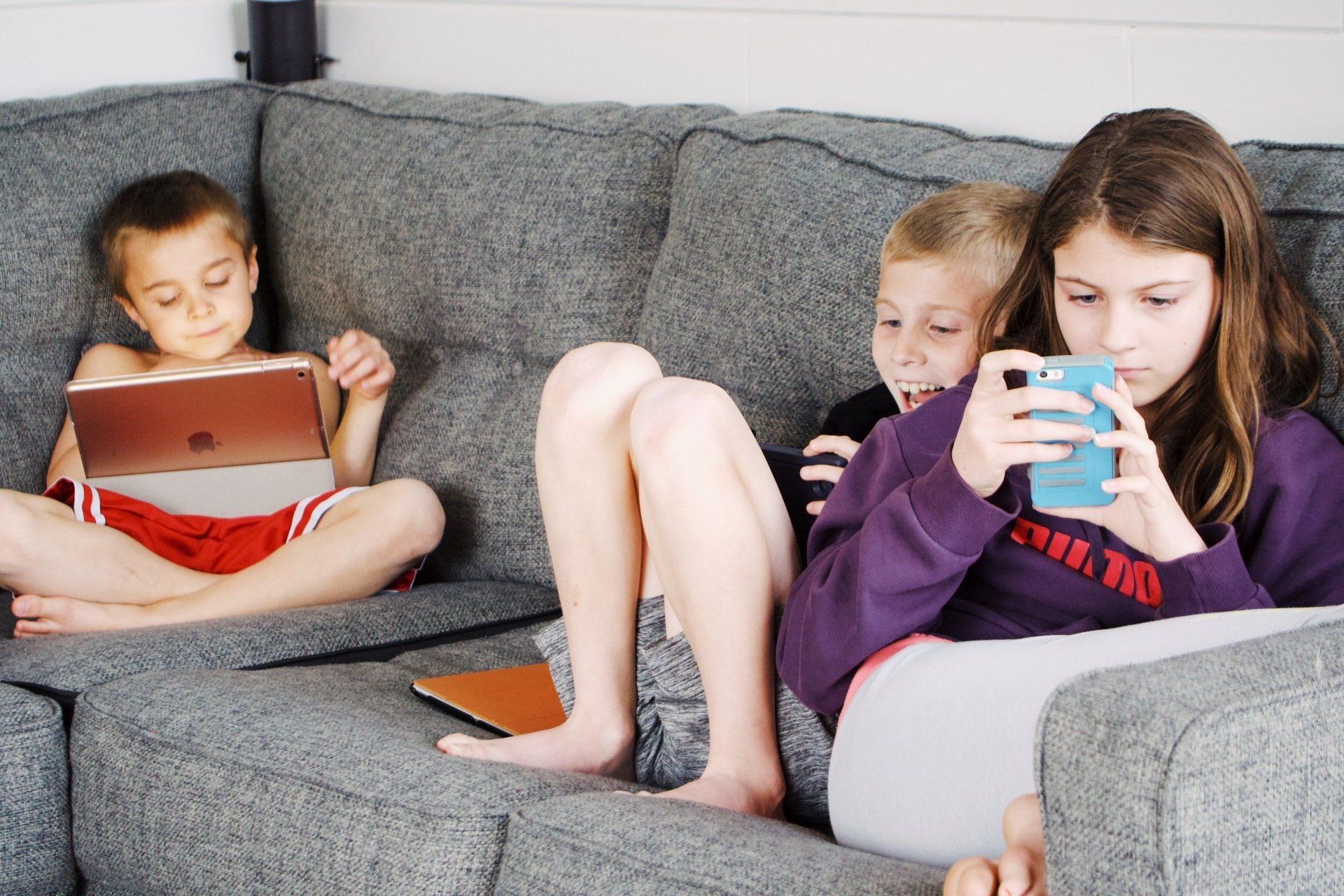 Three children are sitting on a couch looking at their phones.